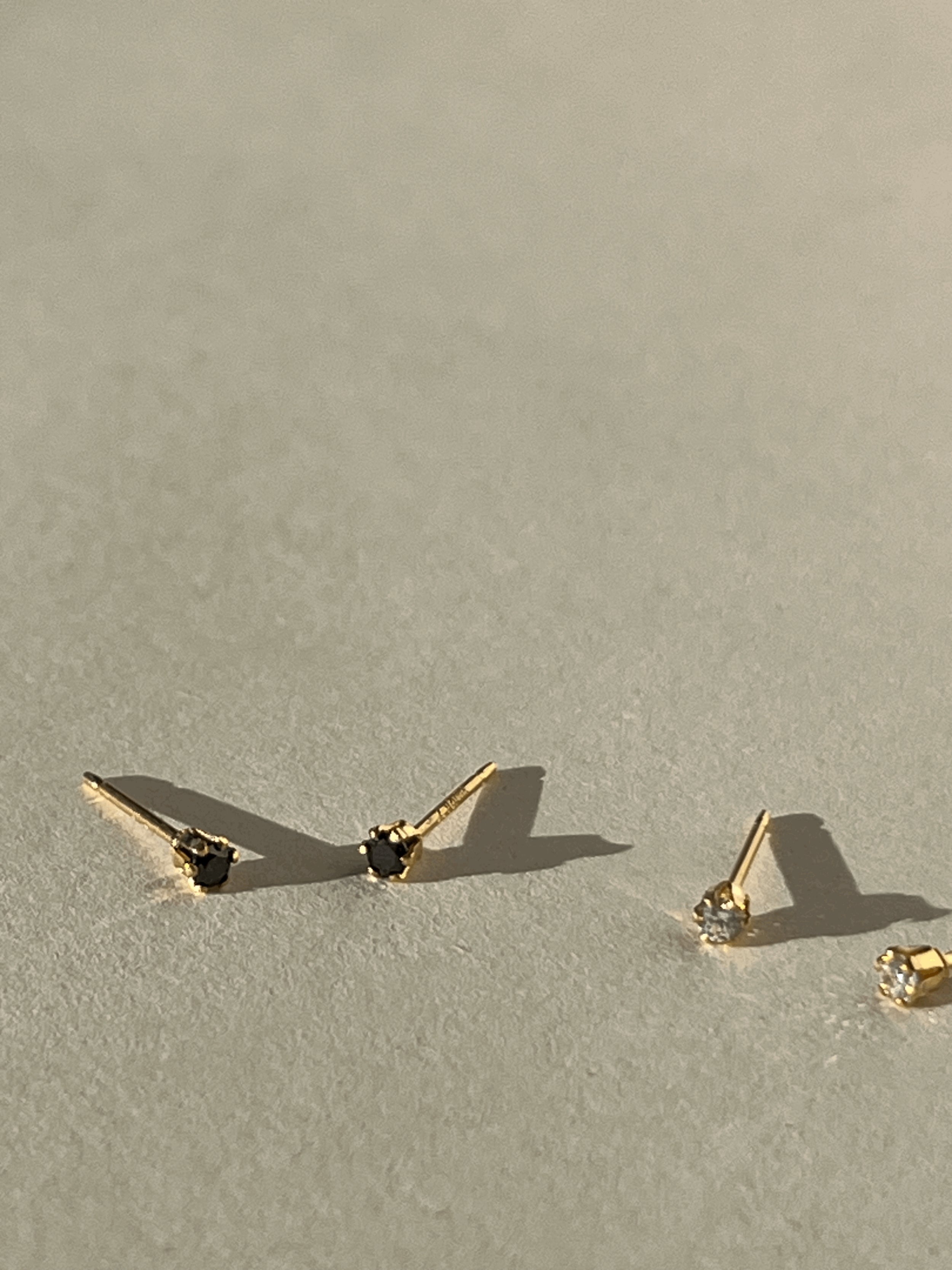Close-up of two pairs of tiny yellow gold-filled cubic zirconia stud earrings, one in black and one in clear, on a neutral background