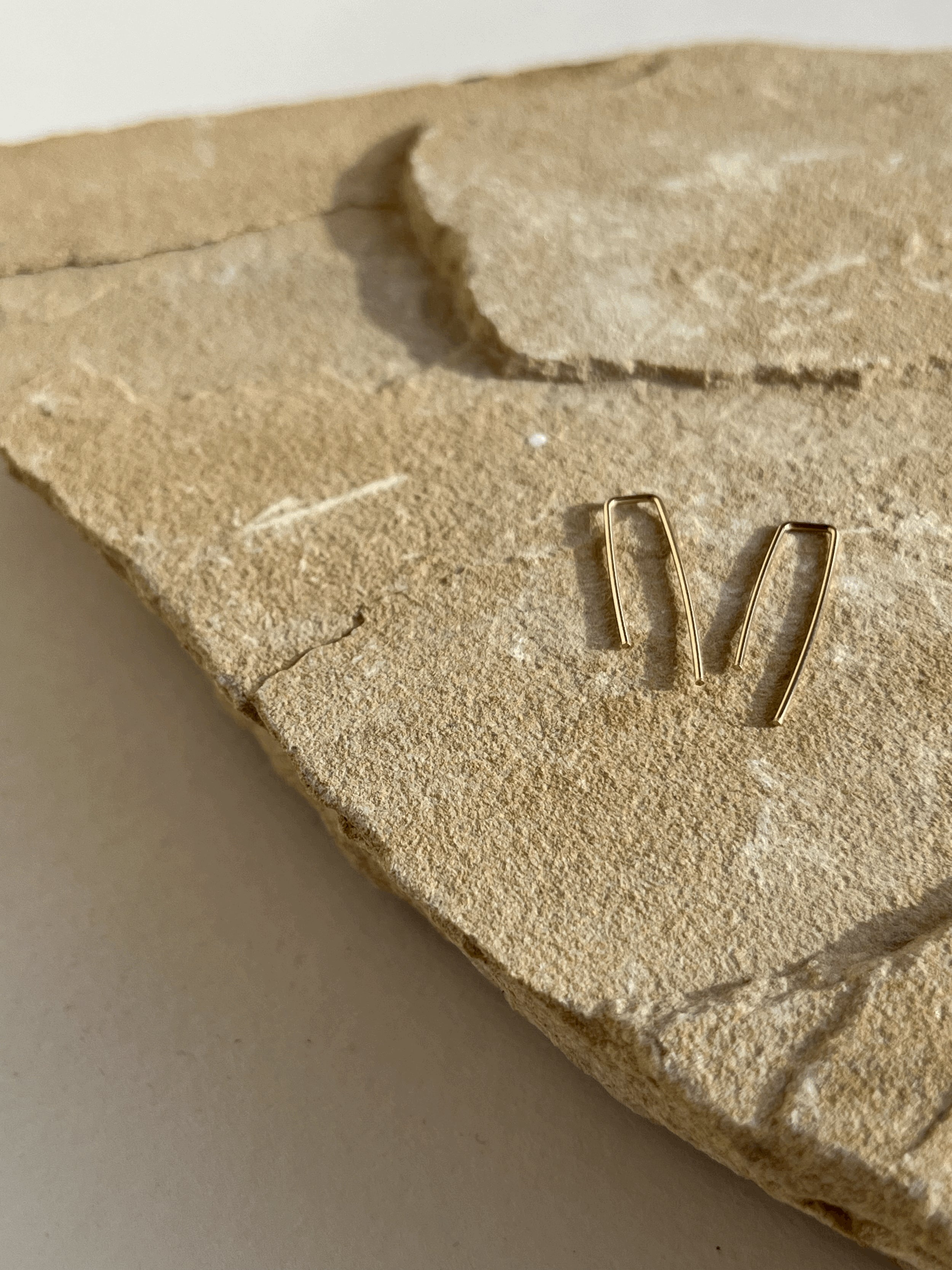 Angled close-up of yellow gold-filled long petite staple earrings resting on a textured rock, capturing the smooth design