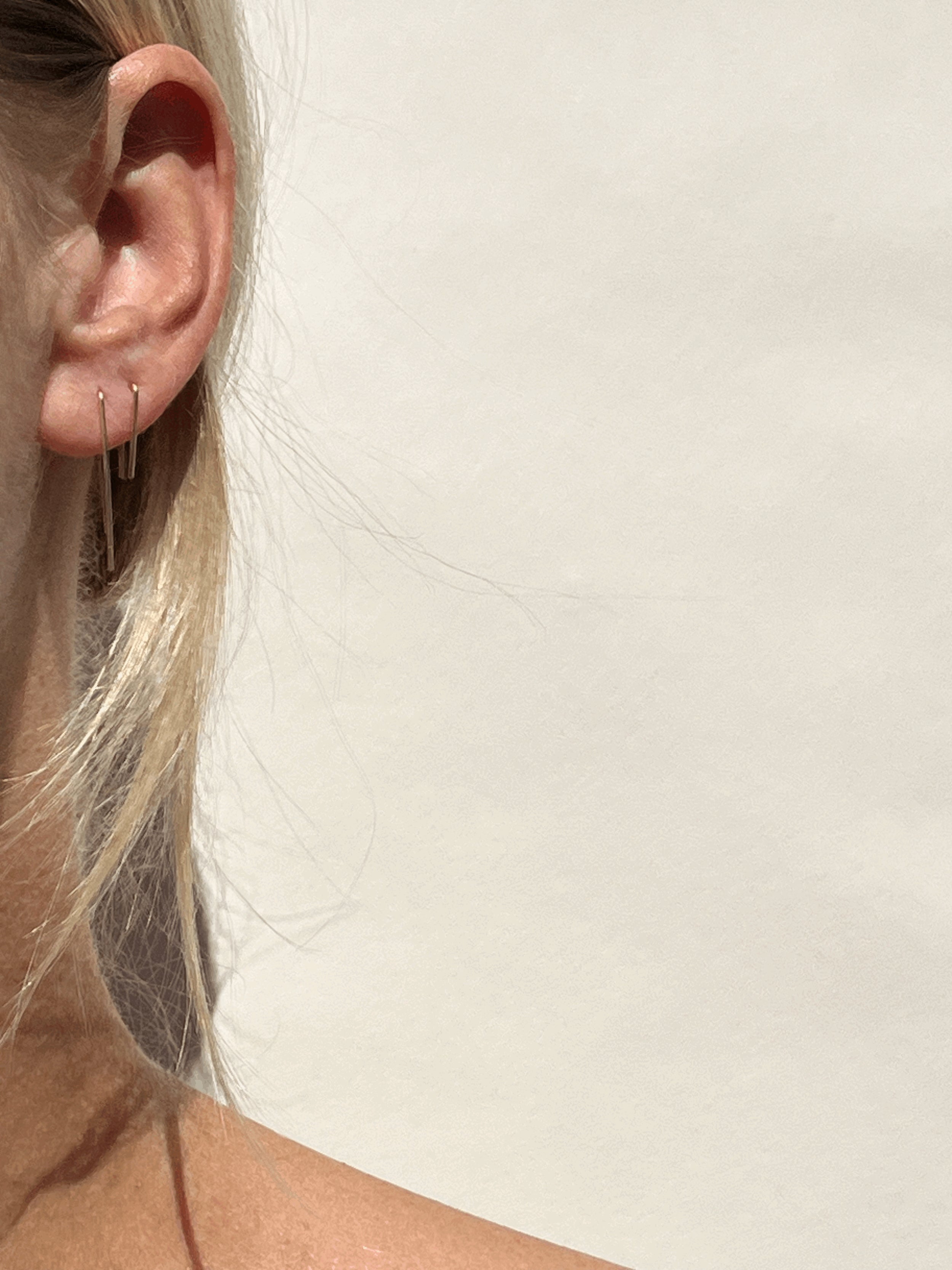 Model wearing long petite staple earrings in yellow gold-filled in her first piercing, with short petite staple earrings in her second piercing, soft focus on ear