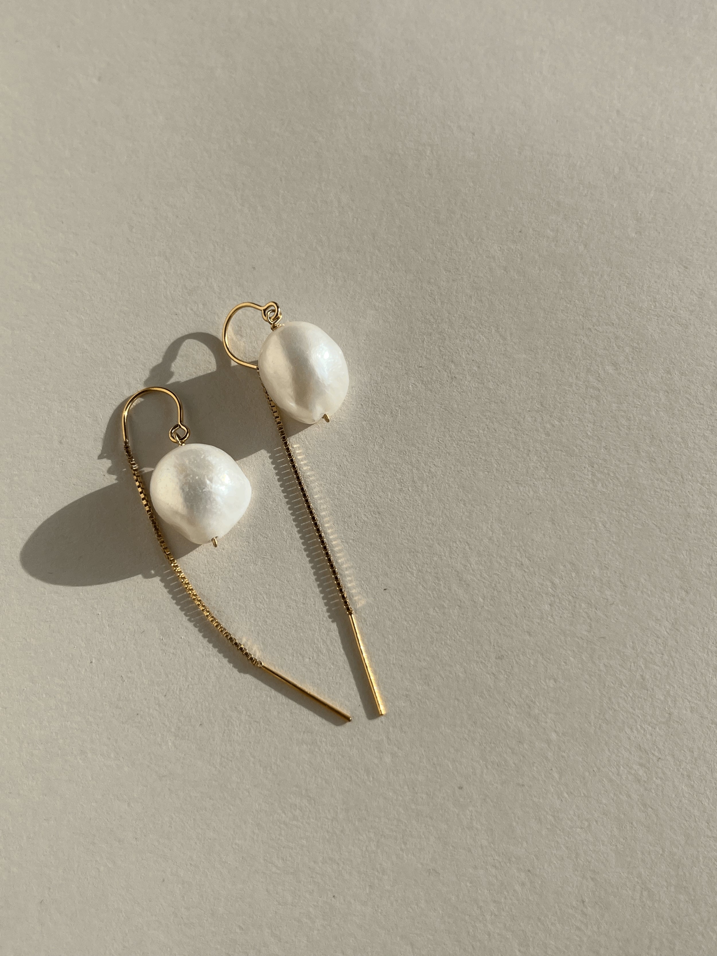 Close-up of the Medium Freshwater Pearl Drop Threader Earrings on a cream backdrop, featuring 12x14mm pearls on minimalist U-threader backs