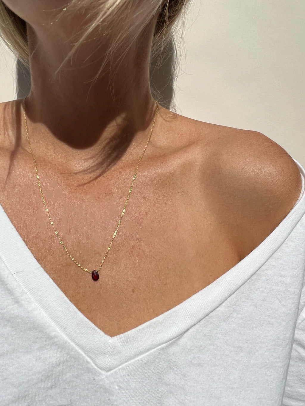 Model wearing minimalist yellow gold-filled garnet drop necklace with natural stone pendant, styled with a white v-neck top