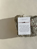My Energy Is Sacred bracelet with deep red cord and gold-filled findings displayed on its printed bracelet card, resting on a rock