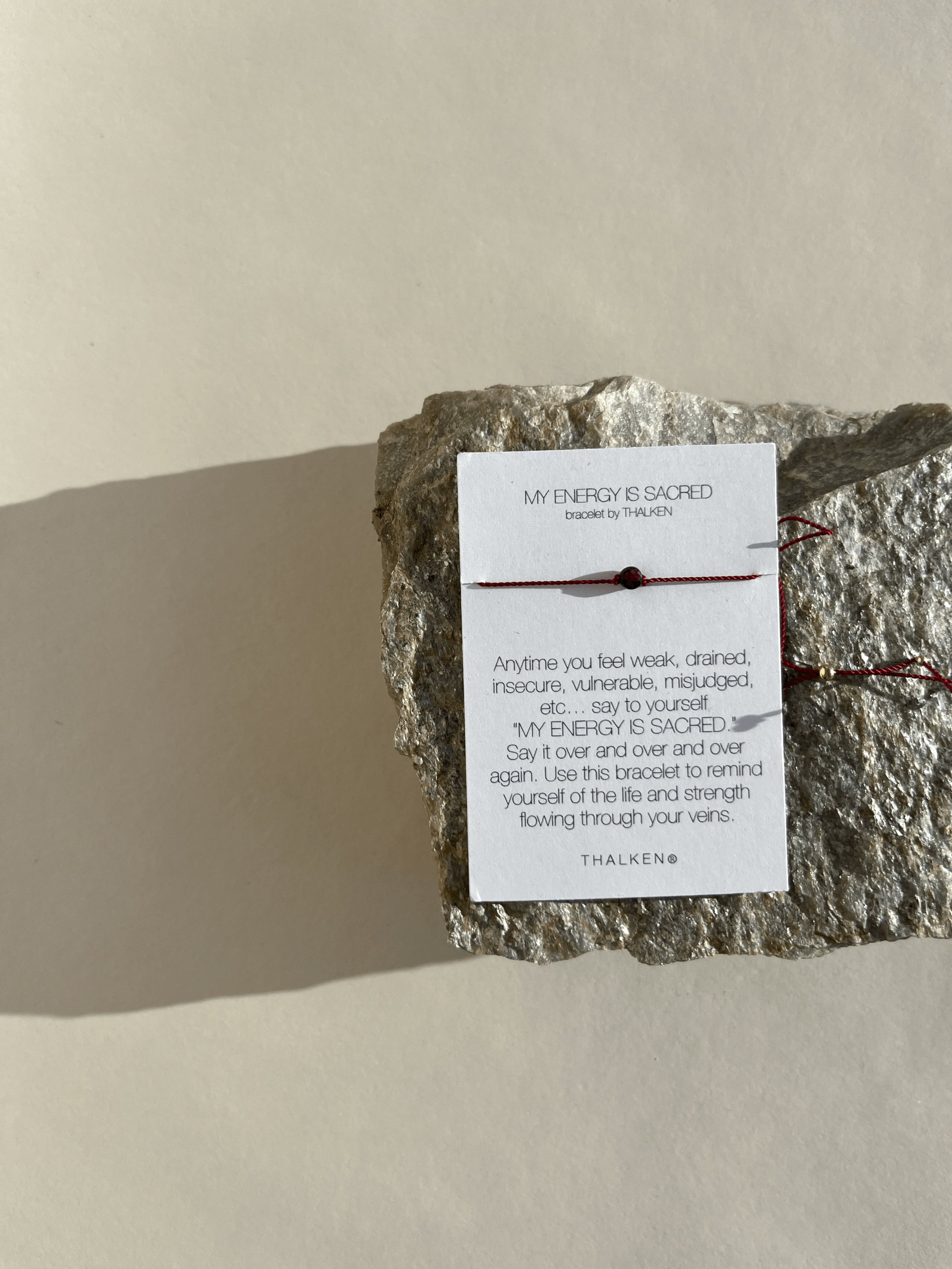 My Energy Is Sacred bracelet with deep red cord and gold-filled findings displayed on its printed bracelet card, resting on a rock