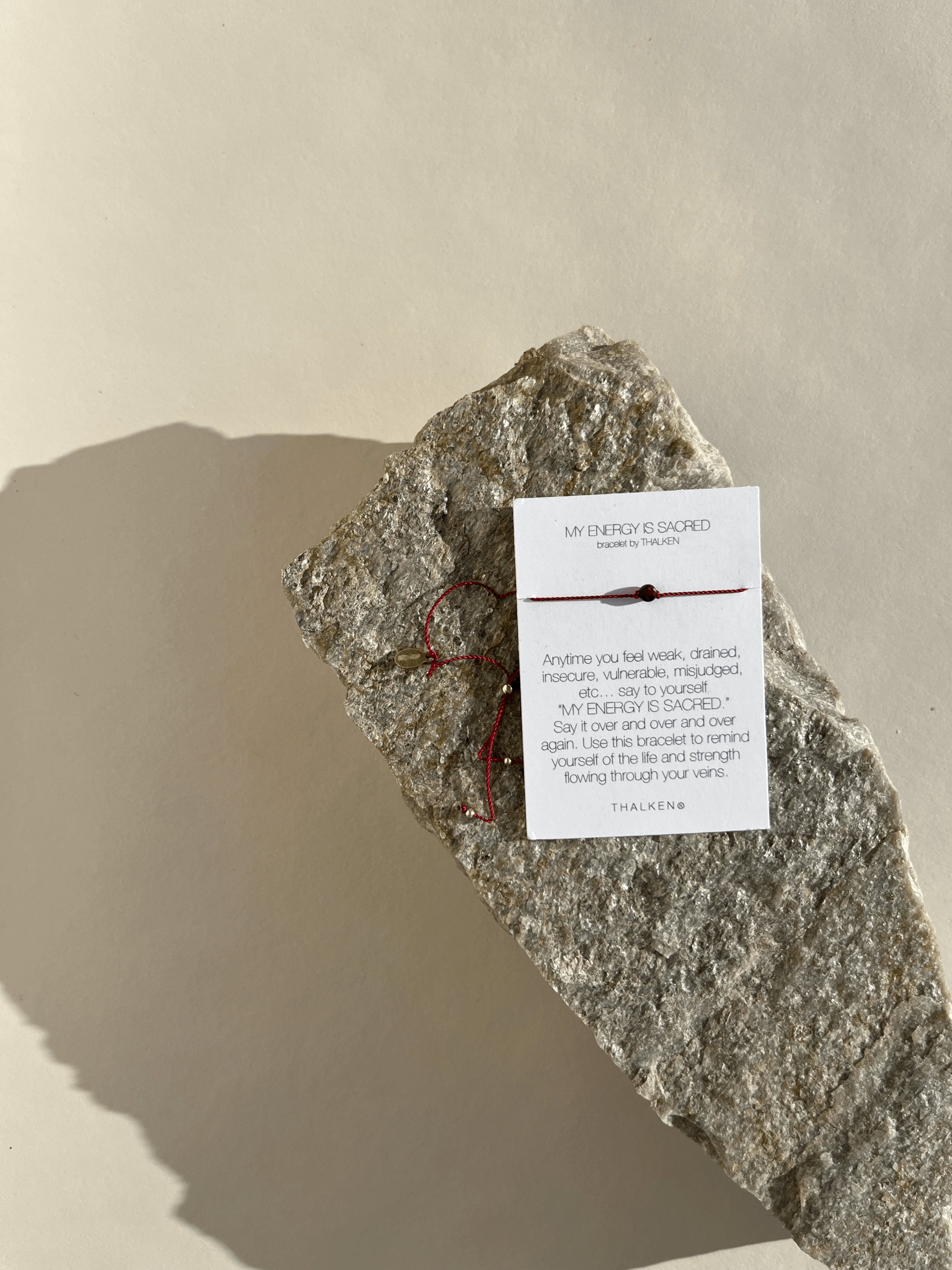 My Energy Is Sacred bracelet with deep red cord and gold-filled findings displayed on its printed bracelet card, resting on a rock from a slightly different angle