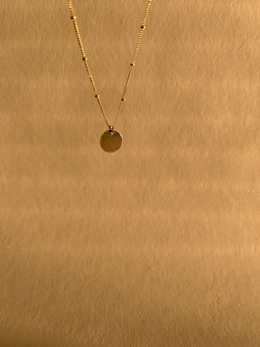 Yellow gold-filled My Favorite Necklace suspended higher in the frame against a warm neutral backdrop, showing its delicate chain, fixed beads, and sliding disc