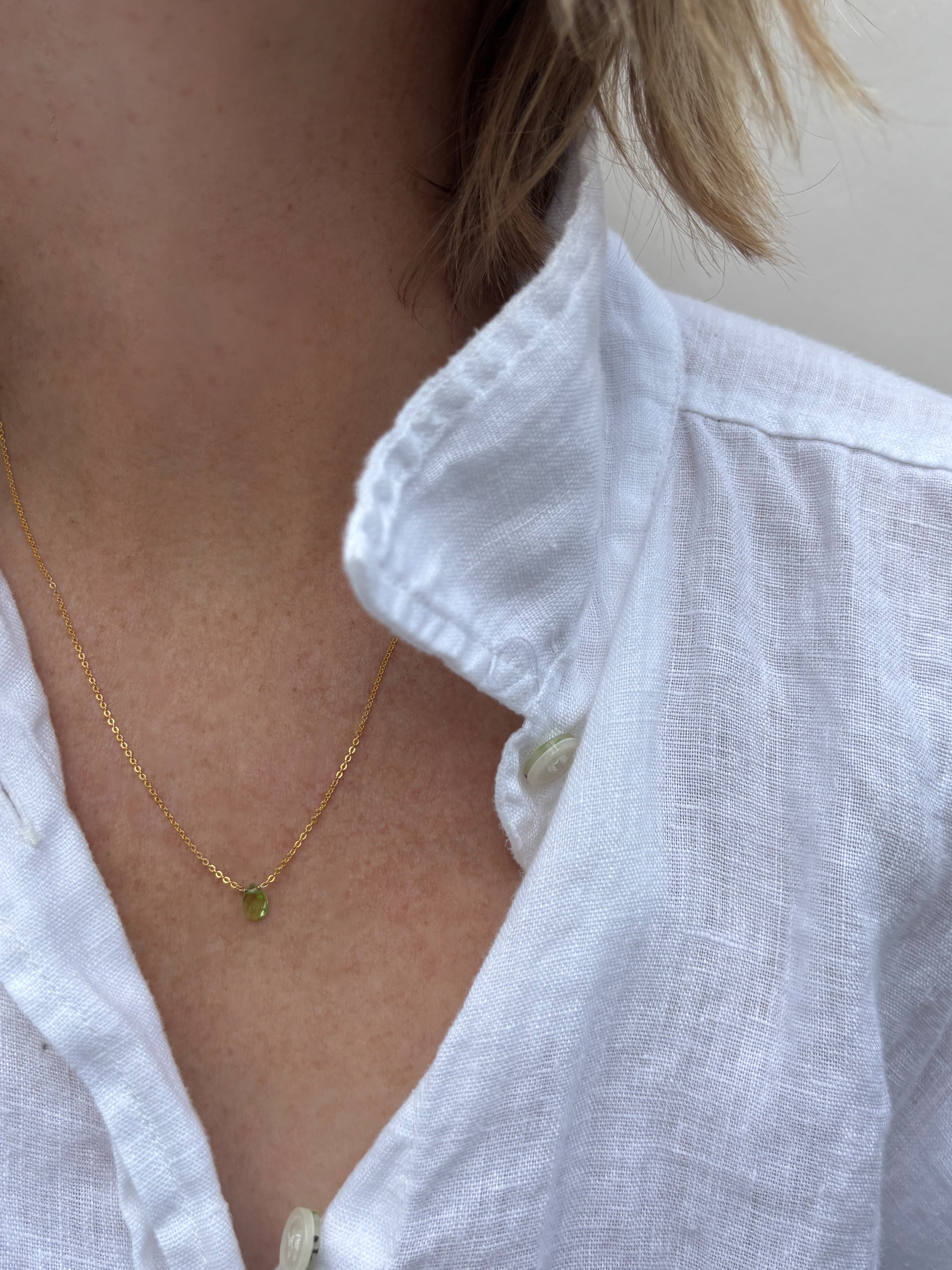 Close-up of woman wearing dainty peridot drop necklace in white linen shirt