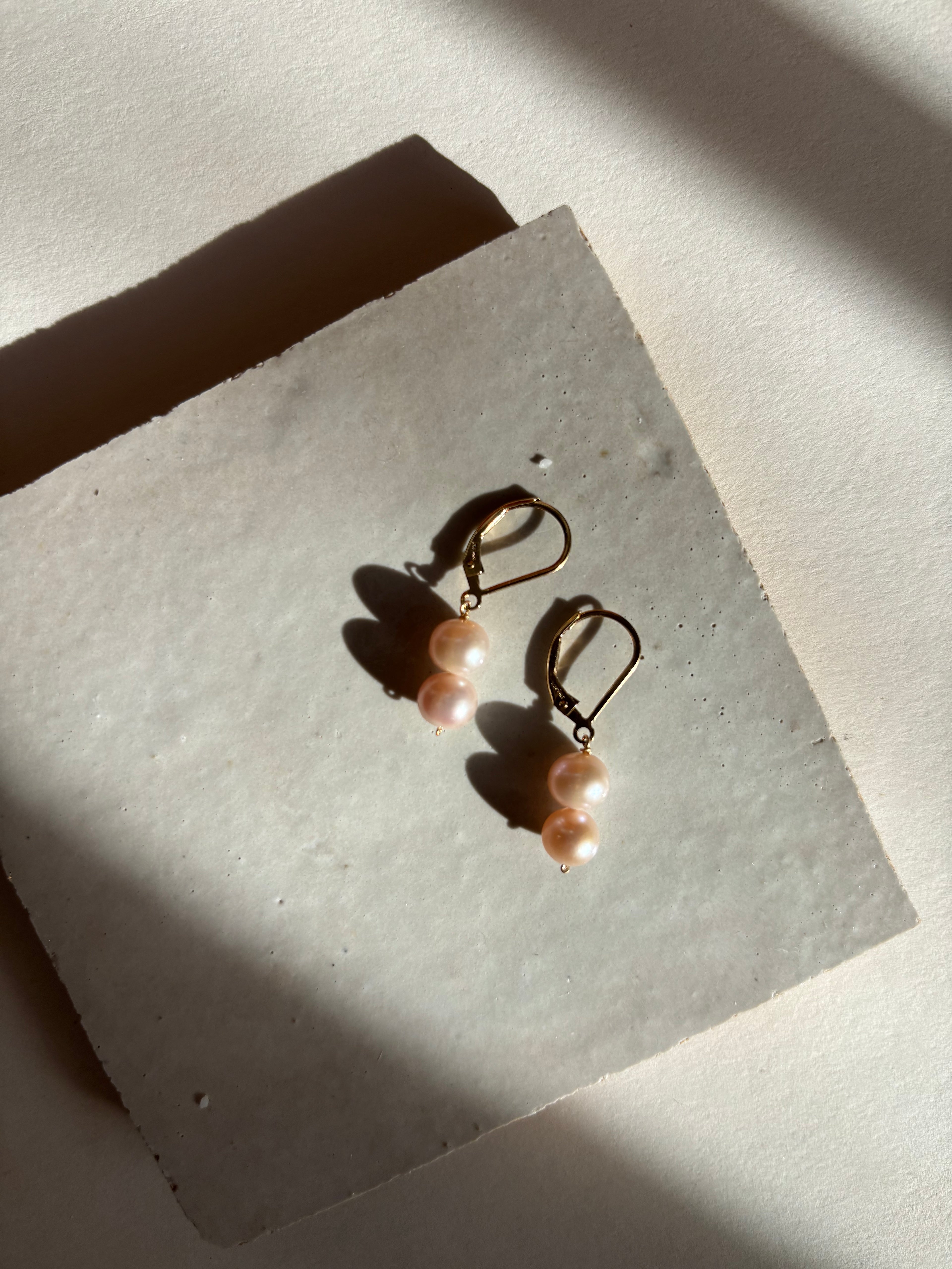 Double pearl drop leverback earrings with blush (soft pink) freshwater pearls shown on an organic surface in warm, moody lighting.