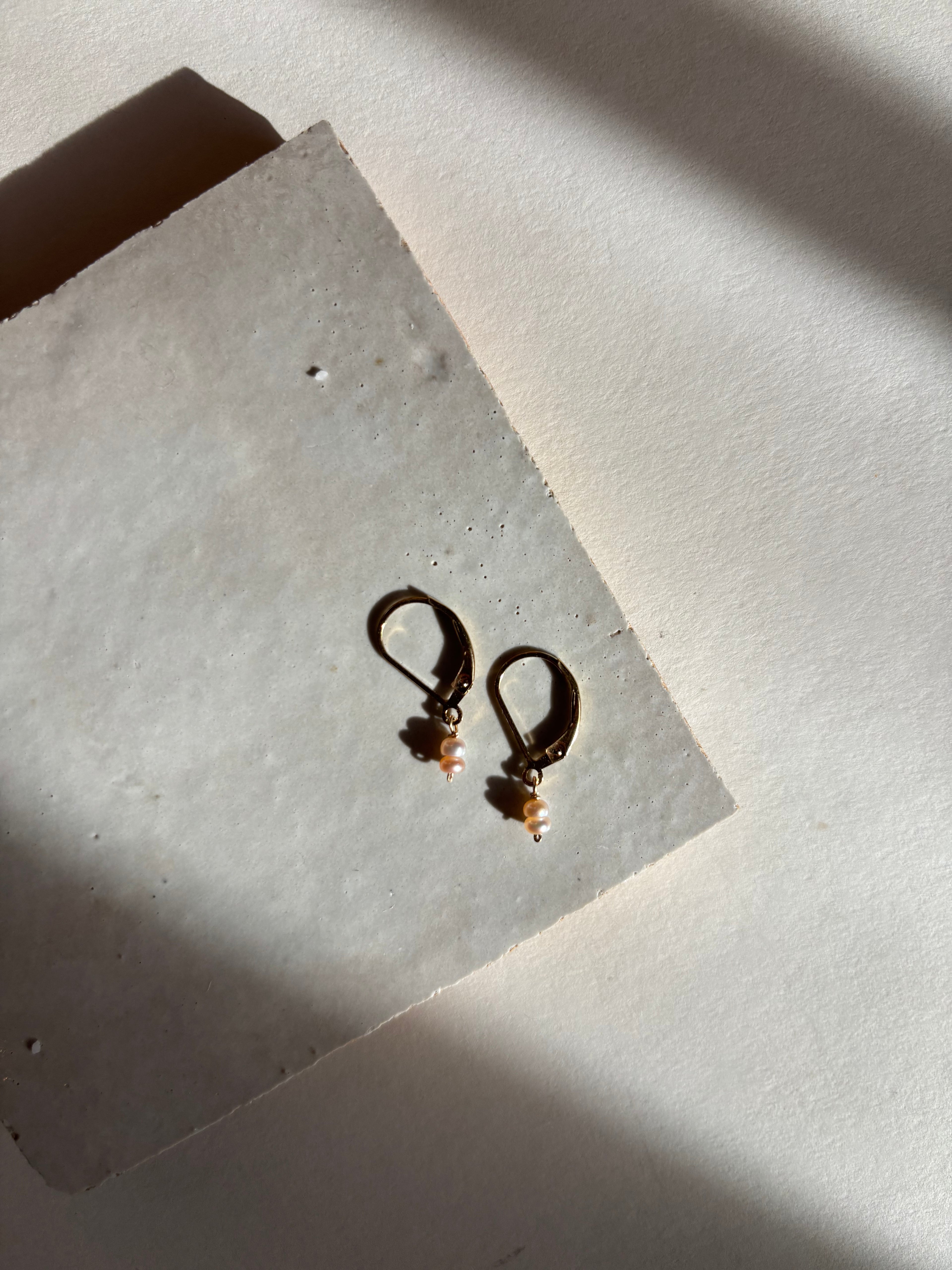 Delicate blush (soft pink) double pearl drop Duet Extra Small earrings on an organic surface with warm, moody lighting showing the full earring design and pearl detail.