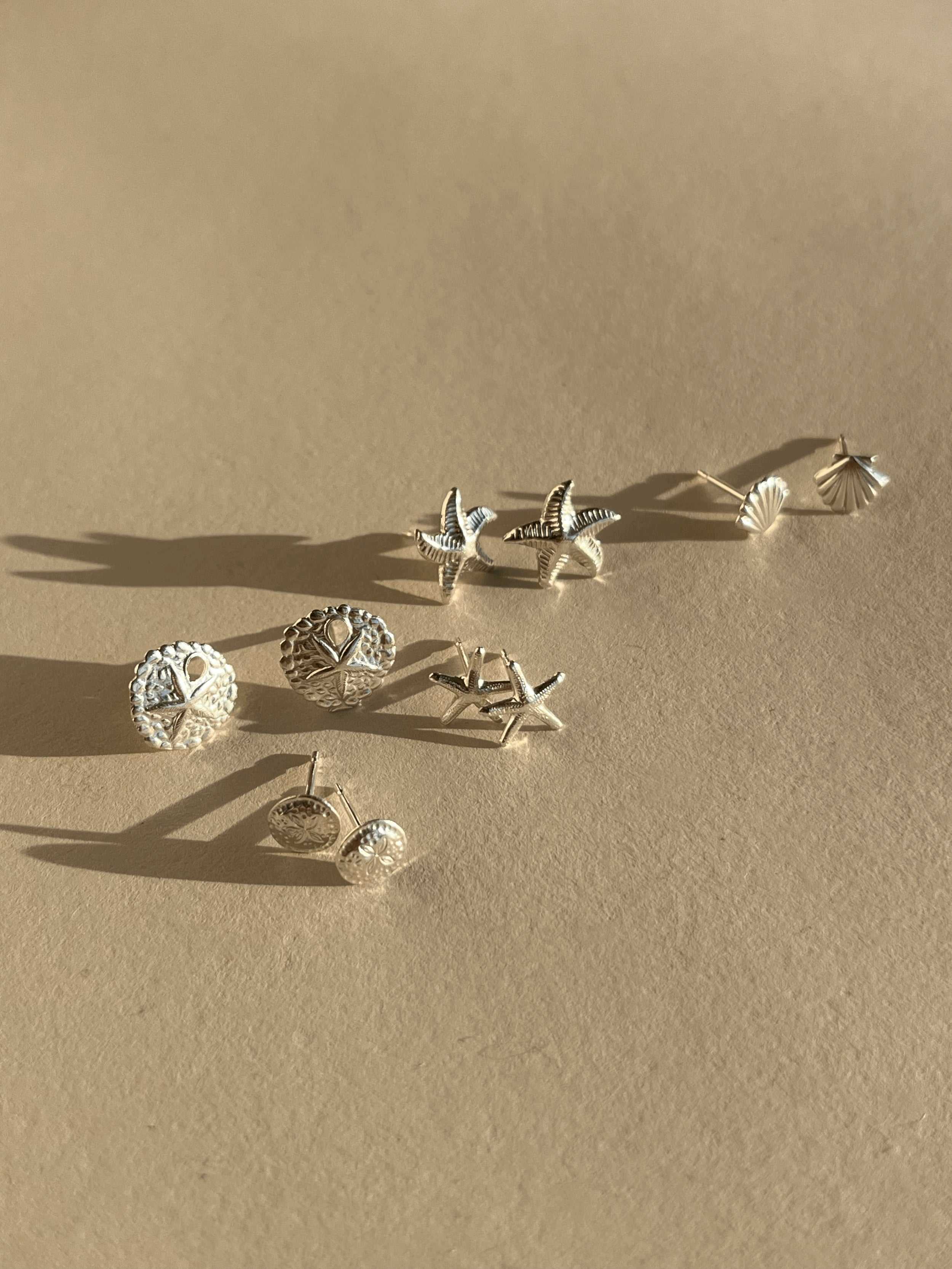 Five pairs of sterling silver sea-inspired stud earrings—sand dollars, starfish, and shells—displayed on a neutral backdrop