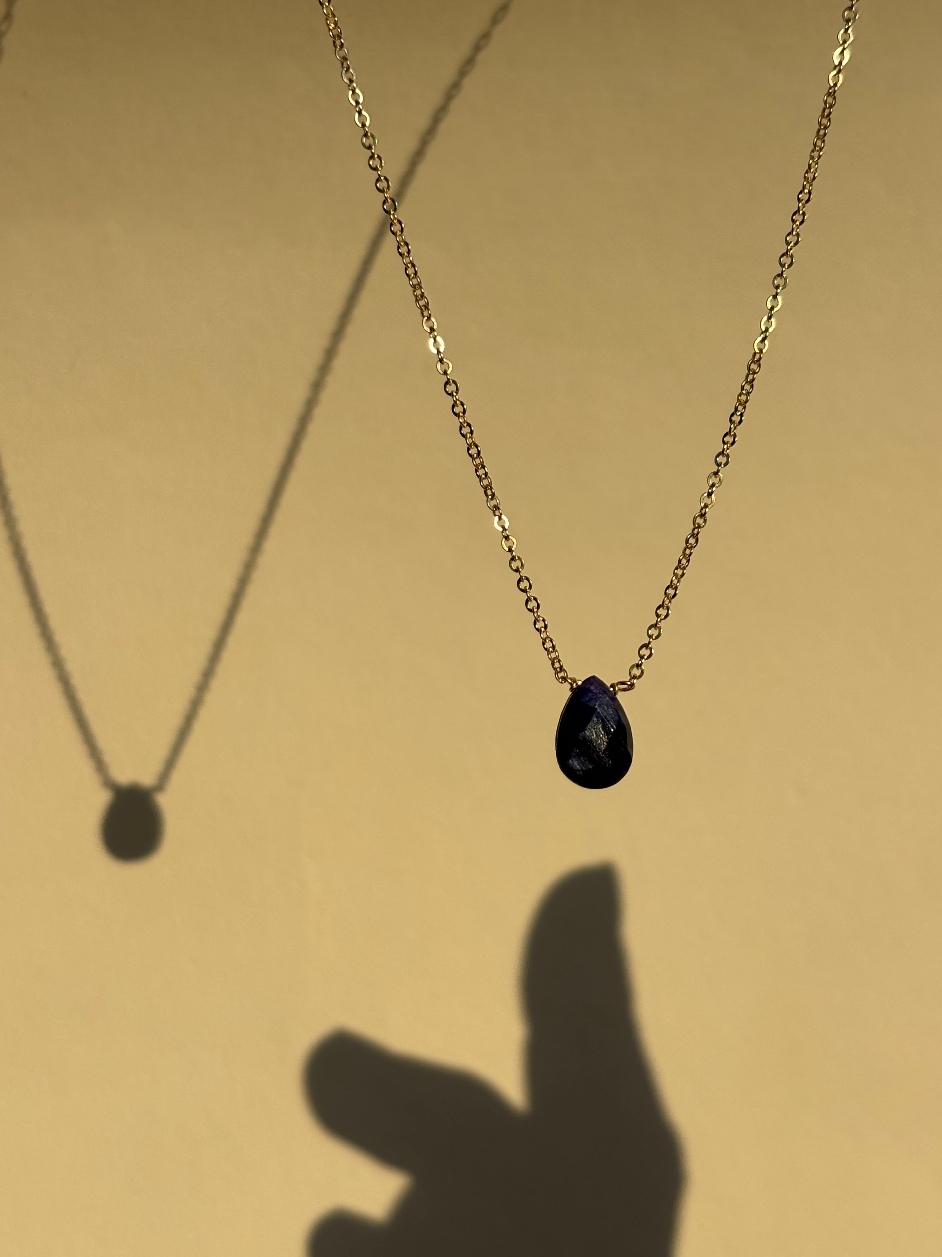 Dark blue natural sapphire stone drop necklace in yellow gold-filled suspended in the air with a shadow of fingers reaching out towards the necklace
