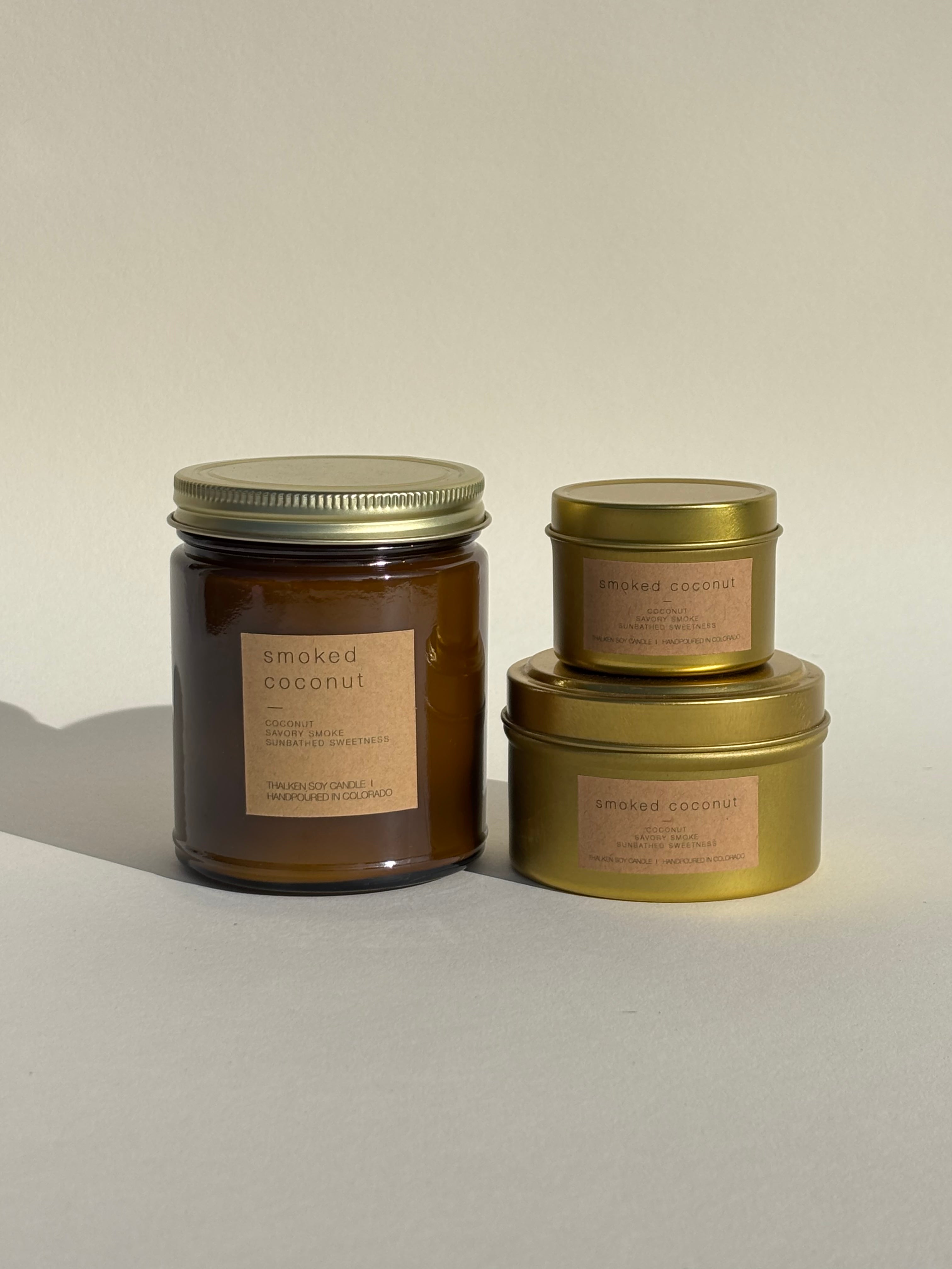 Photo of Thalken’s Smoked Coconut candle shown in three vessel options — sleek gold tins (2oz and 6oz) and a soft-glow glass jar (8oz). Earthy scent profile with notes of coconut, savory smoke, and sunbathed sweetness.