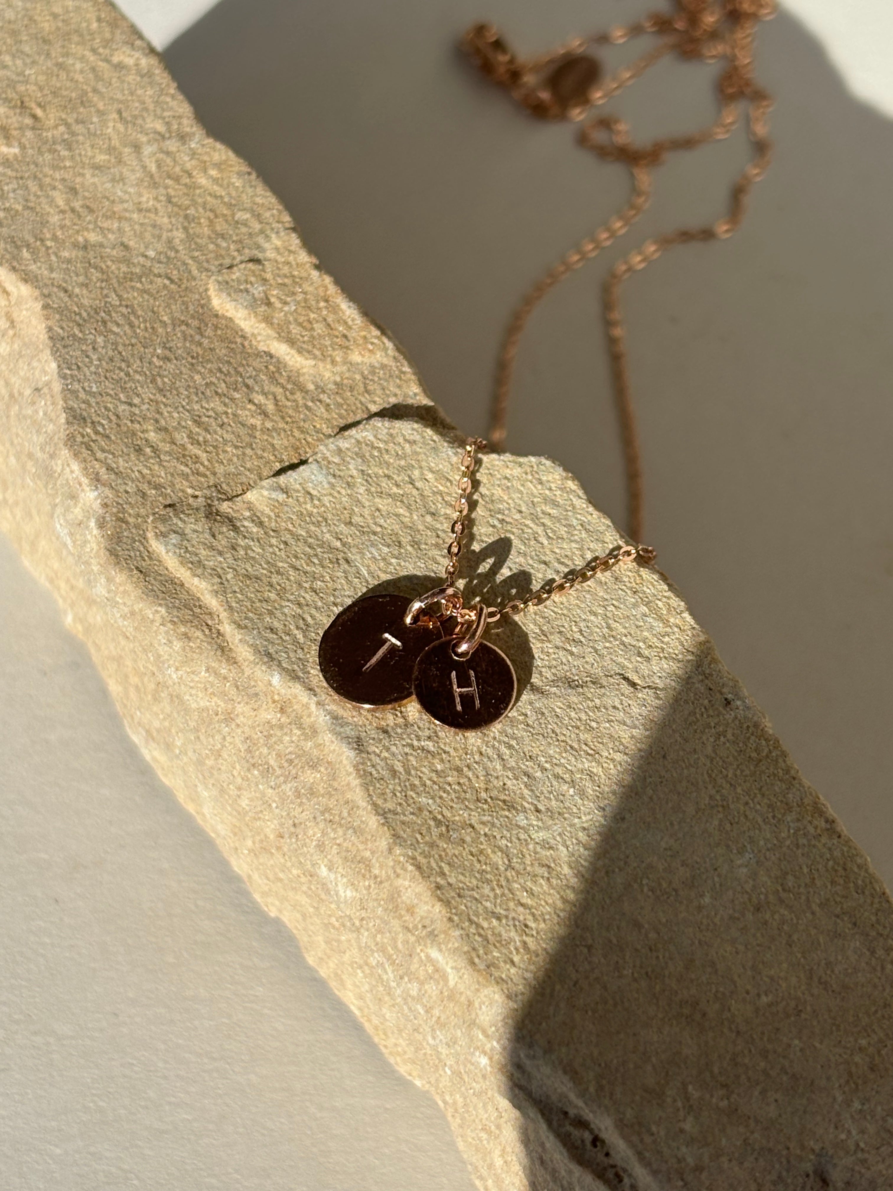 Rose gold-filled 7mm and 9mm stamped letter disc charms with “H” and “T” laying on a natural rock surface