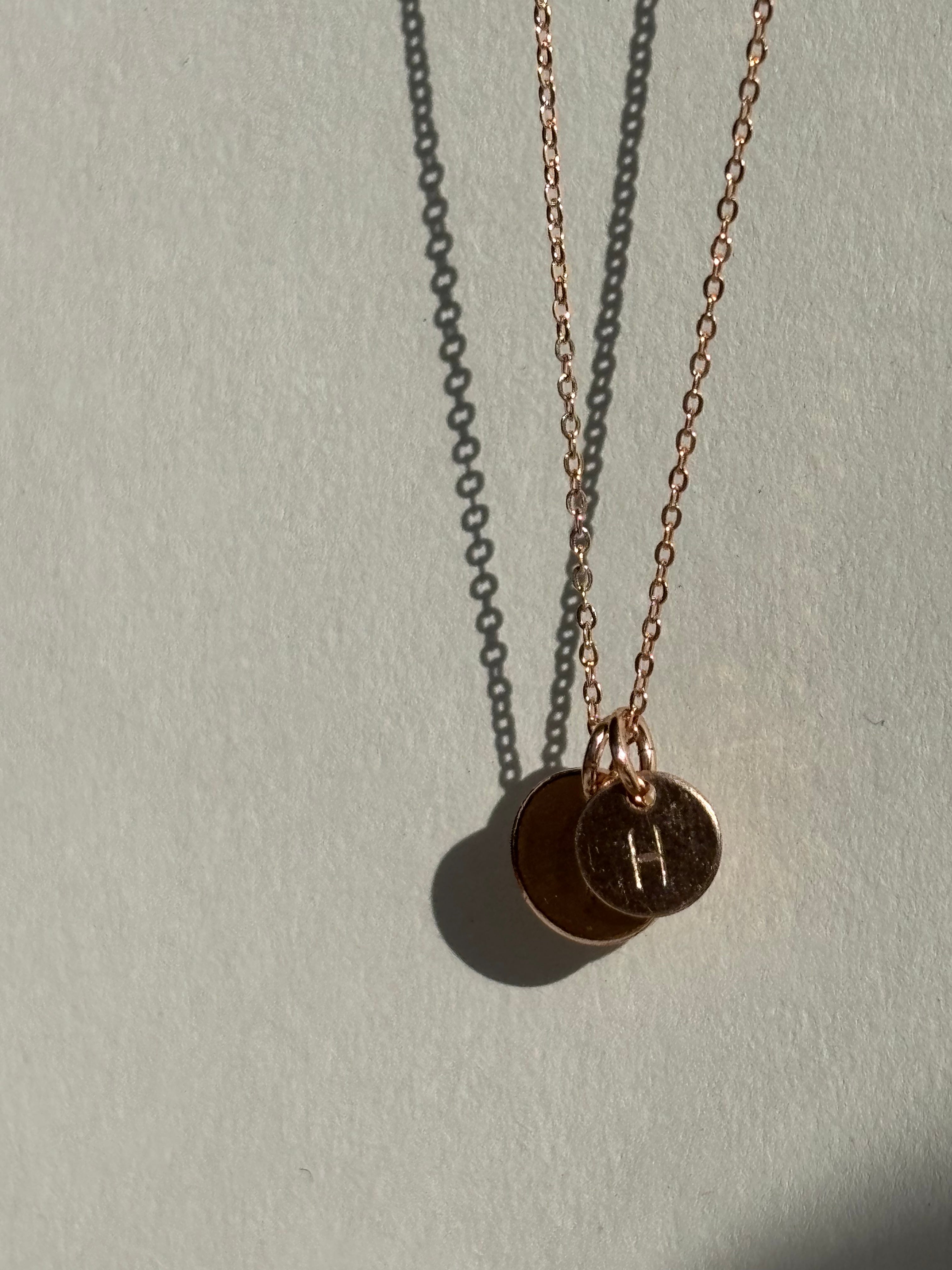 Close-up of a rose gold-filled necklace with two charms: a small 7mm disc with an uppercase “H” in front and a medium 9mm disc with a “T” behind it