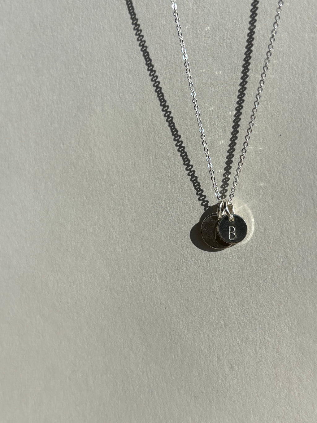 Alternate angle of a sterling silver 7mm “B” disc charm in front of a 9mm “H” disc charm on a chain