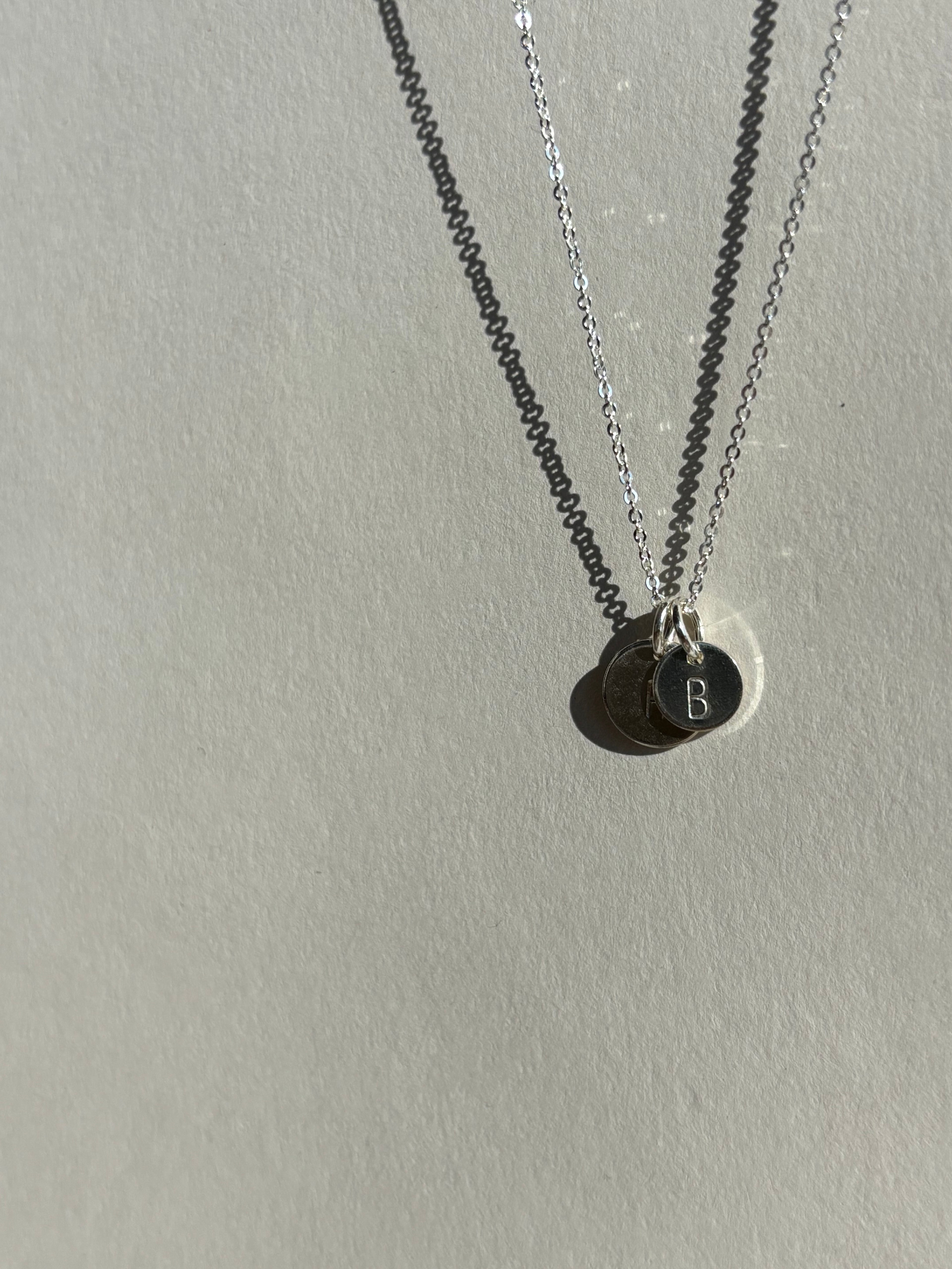 Alternate angle of a sterling silver 7mm “B” disc charm in front of a 9mm “H” disc charm on a chain