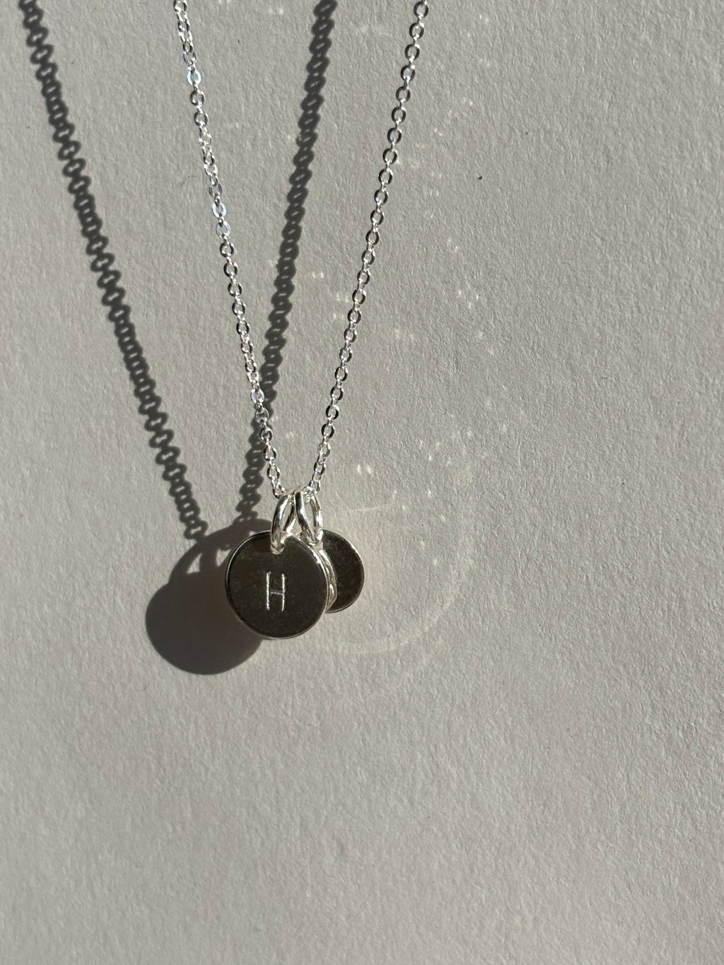 Close-up of a sterling silver 9mm “H” disc charm in front of a 7mm “B” disc charm on a chain