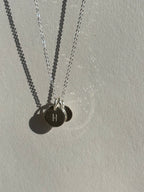 Close-up of a sterling silver 9mm “H” disc charm in front of a 7mm “B” disc charm on a chain