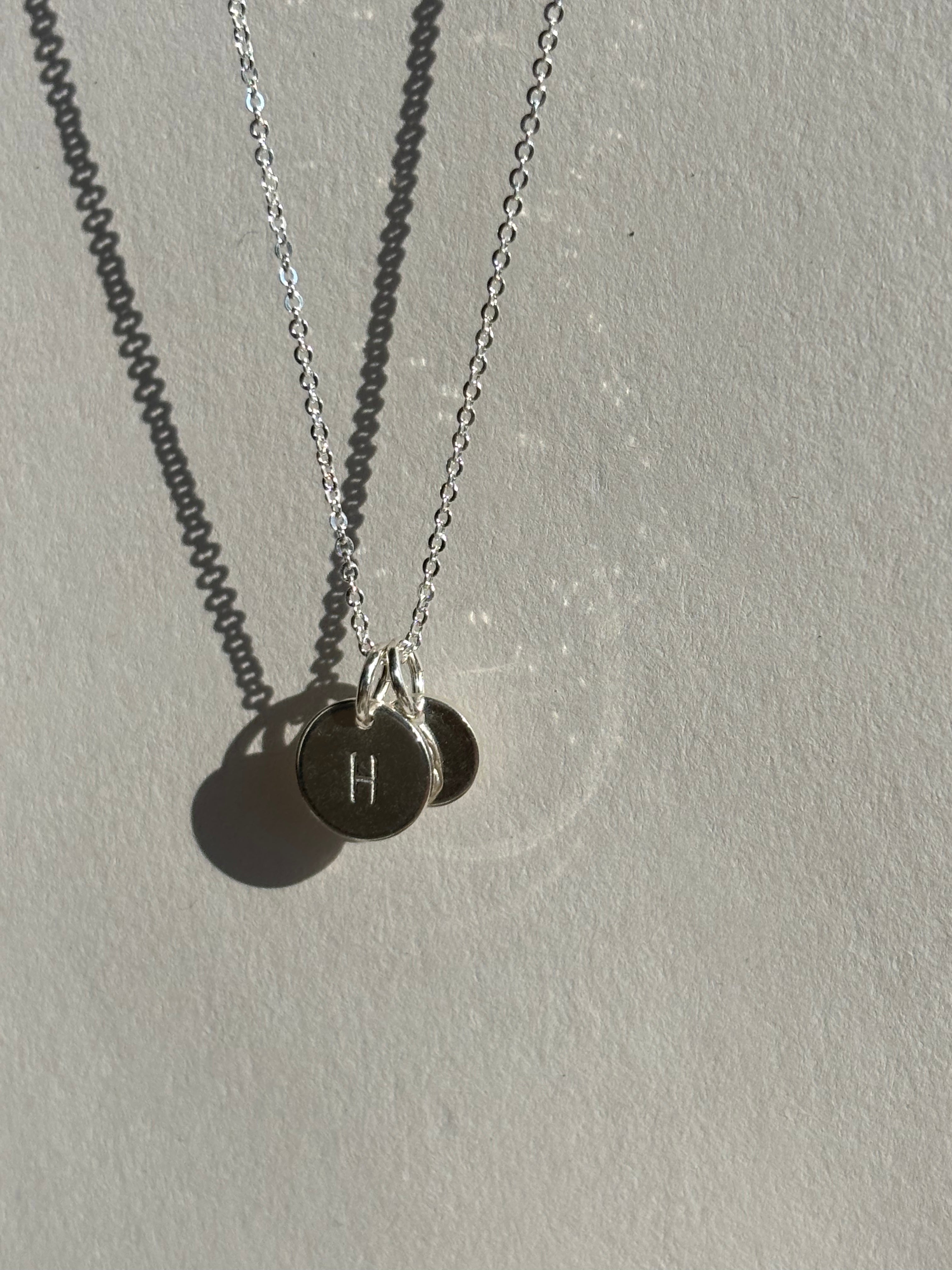 Close-up of a sterling silver 9mm “H” disc charm in front of a 7mm “B” disc charm on a chain