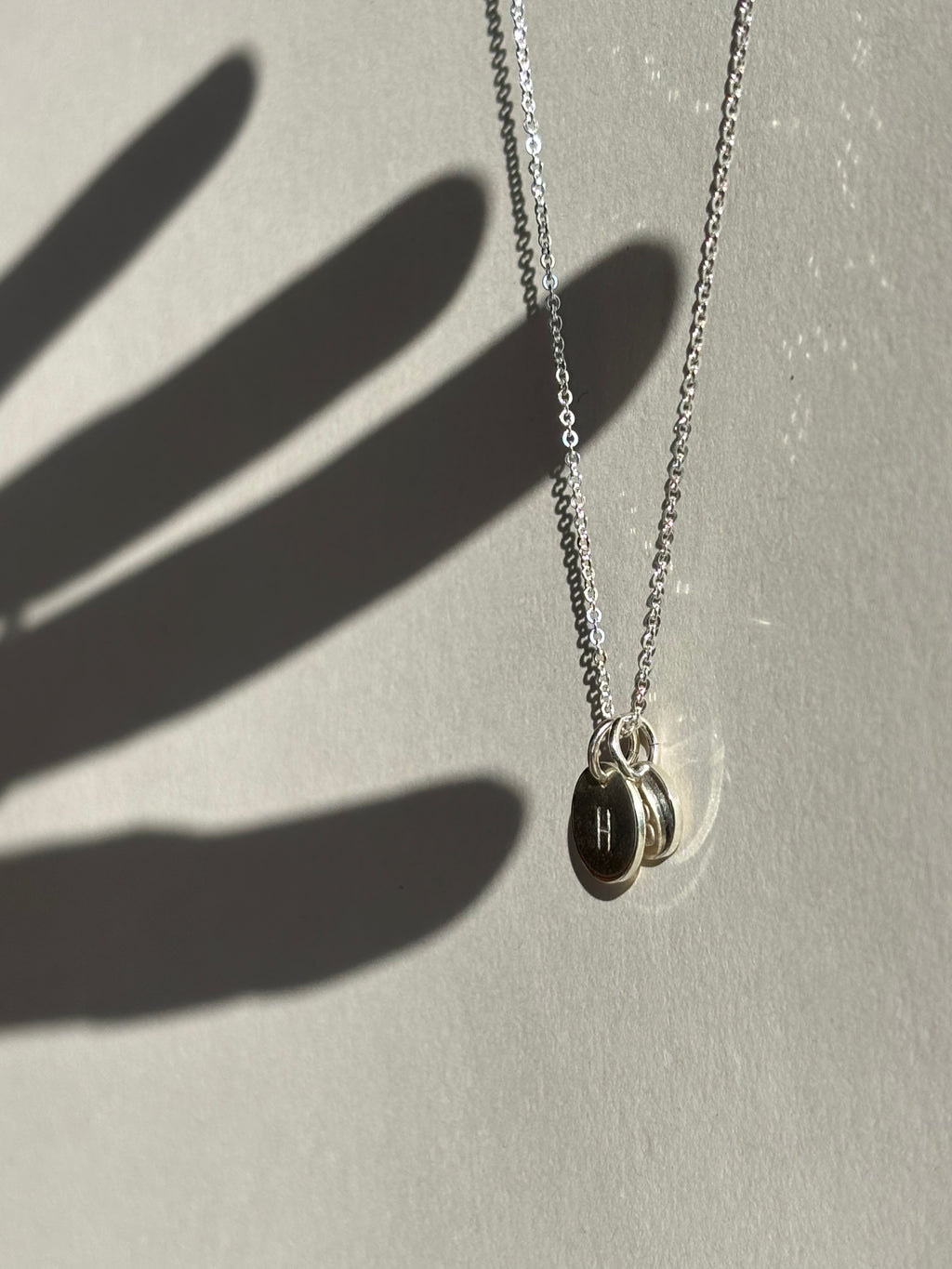 Sterling silver necklace with a 9mm disc charm stamped with “H” layered in front of a 7mm “B” disc, with a hand shadow appearing to touch the charms