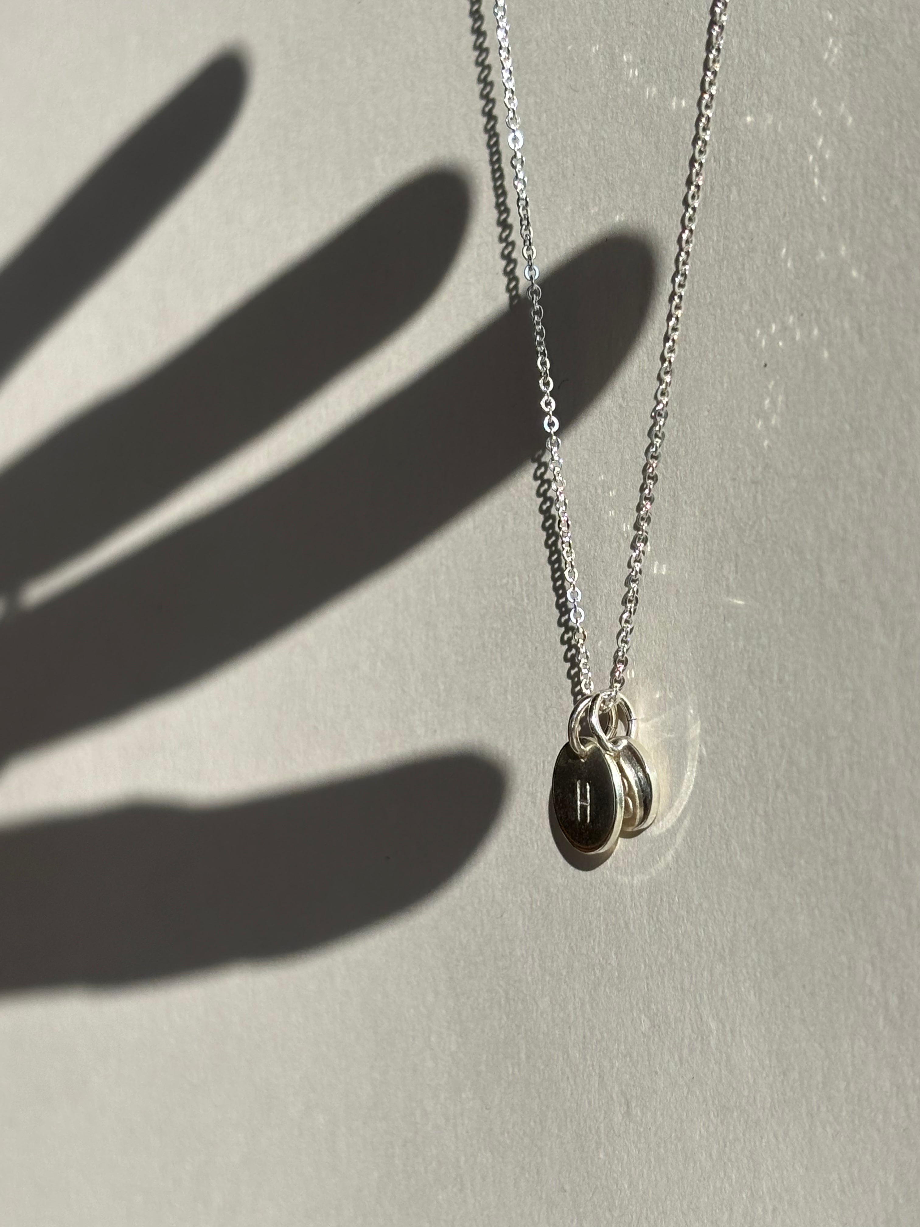 Sterling silver necklace with a 9mm disc charm stamped with “H” layered in front of a 7mm “B” disc, with a hand shadow appearing to touch the charms