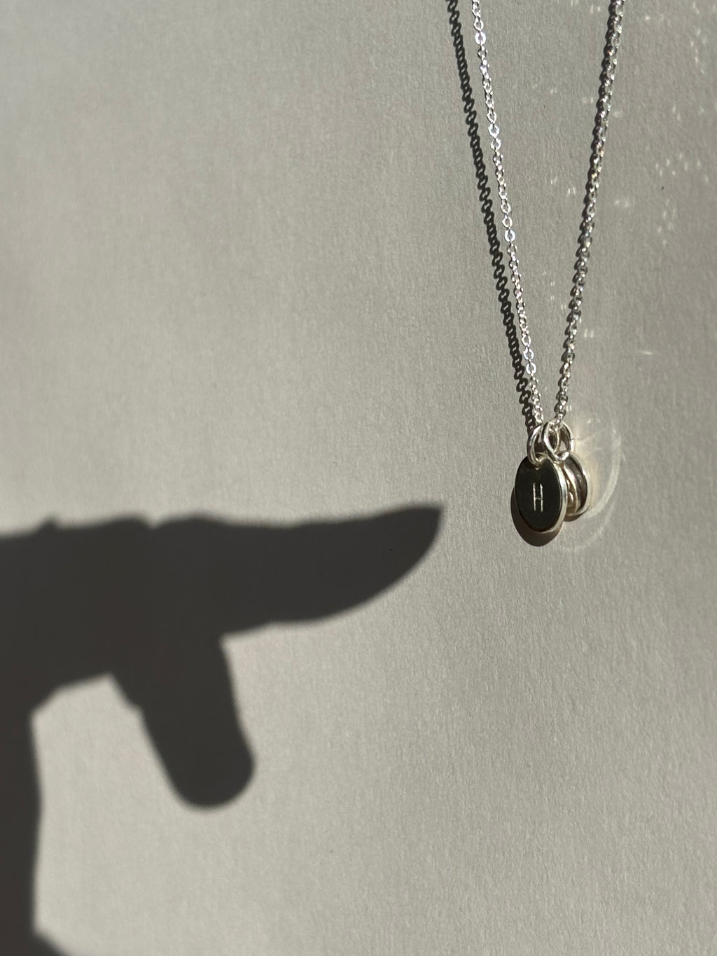 Different angle of a sterling silver necklace with a 9mm “H” disc in front of a 7mm “B” disc charm, with a hand shadow pointing at the charms
