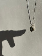 Different angle of a sterling silver necklace with a 9mm “H” disc in front of a 7mm “B” disc charm, with a hand shadow pointing at the charms