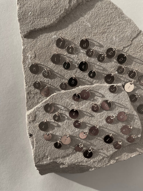 Closer view of sterling silver 7mm and 9mm disc charms with all letters of the alphabet stamped, shown grouped on a light background