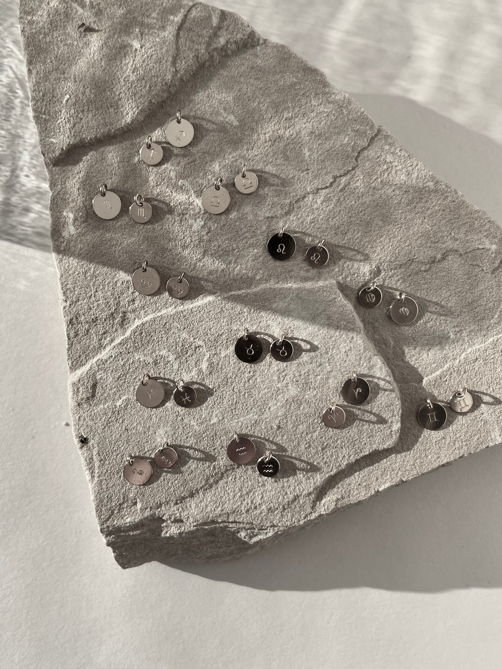 Black and white image of sterling silver 7mm and 9mm disc charms stamped with all zodiac signs, displayed together on a light background