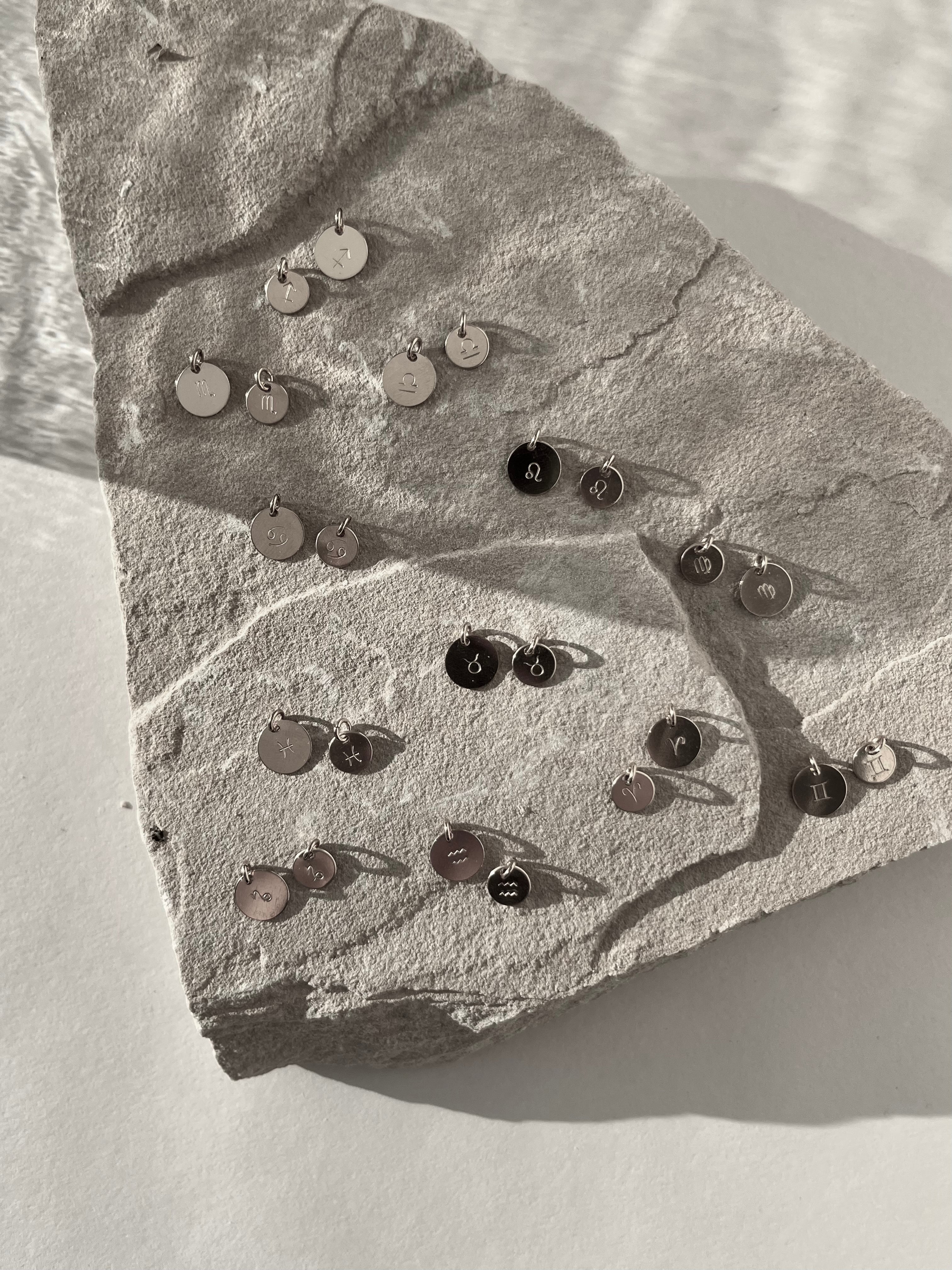 Black and white image of sterling silver 7mm and 9mm disc charms stamped with all zodiac signs, displayed together on a light background