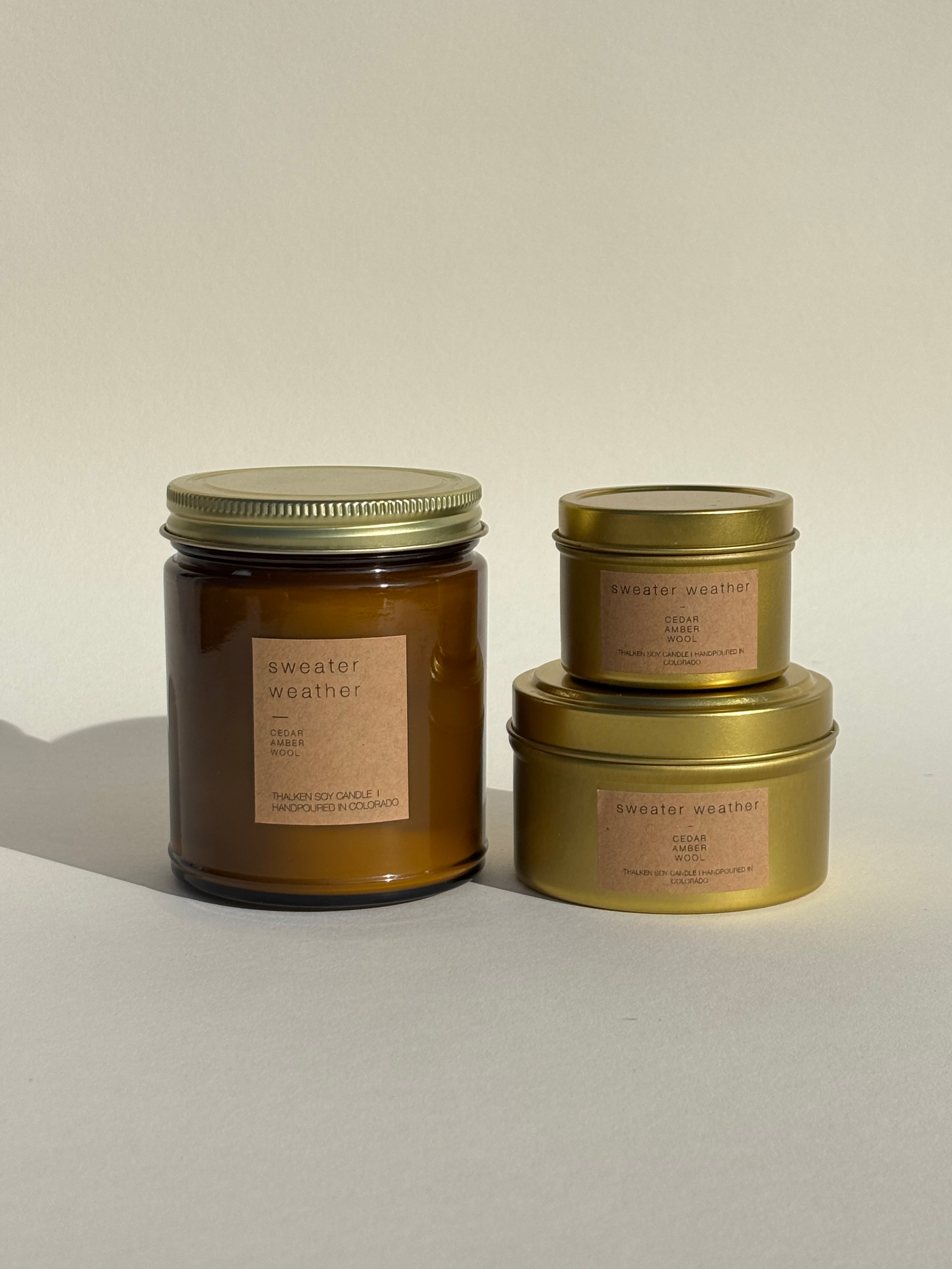 Photo of Thalken’s Sweater Weather candle shown in three vessel options — sleek gold tins (2oz and 6oz) and a soft-glow glass jar (8oz). Earthy scent profile with notes of cedar, amber, and wool.