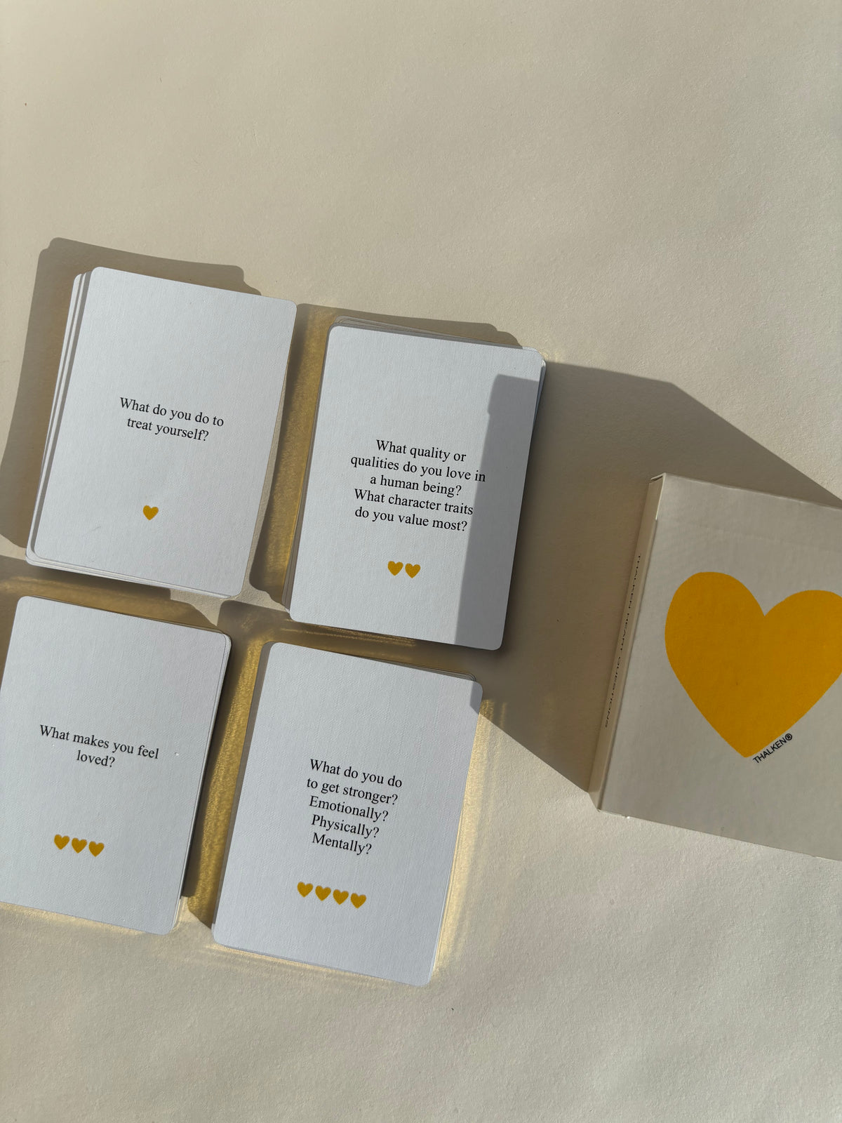Four stacks of Thalken Heart Questions cards, each marked with a different number of hearts to show levels of depth—more hearts mean deeper questions. Beside the stacks lies the deck’s yellow heart container with the Thalken® mark
