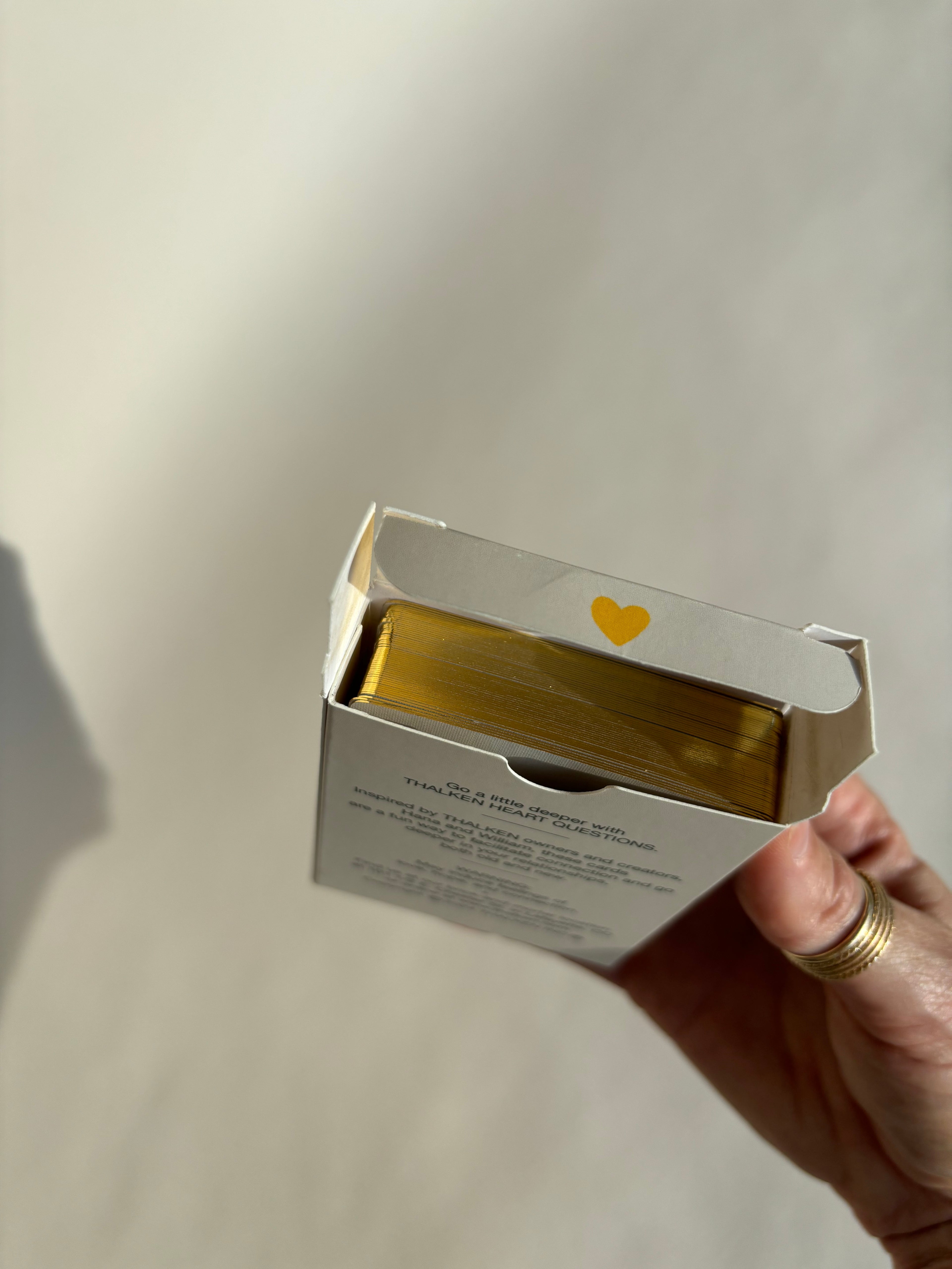 Hand holding the Thalken Heart Questions deck inside its case with a yellow heart, showing both the case design and the gold-gilded edging