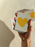 Hand holding a Thalken Heart Questions deck with a large yellow heart on the front and gold-gilded edging, glowing in warm light