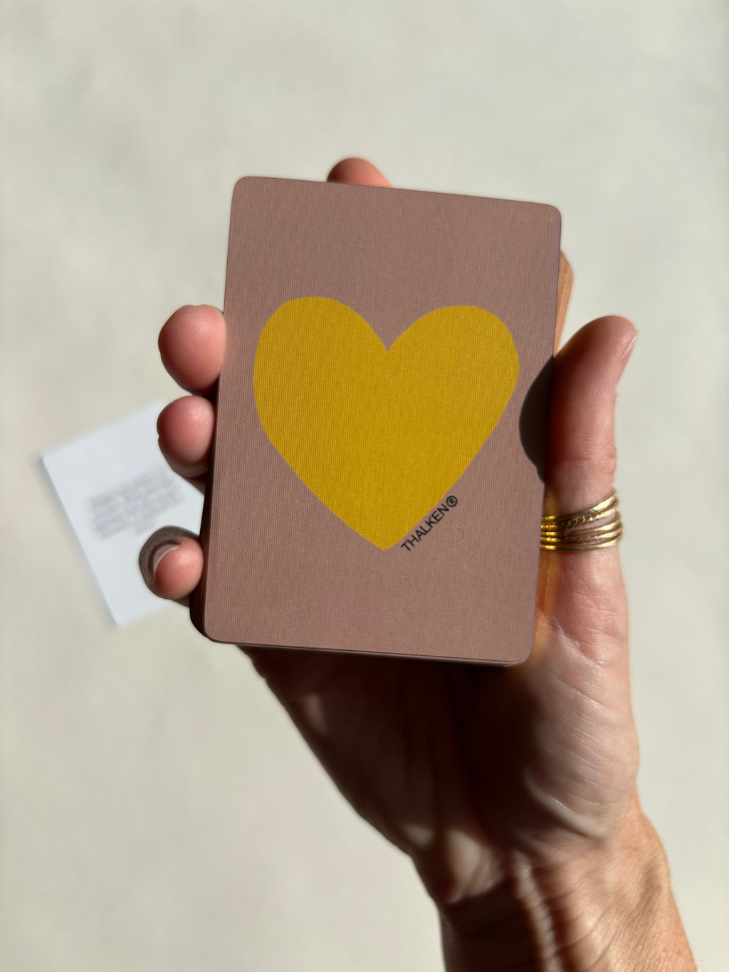 Hand holding Thalken Heart Questions Volume 2 deck with gold-gilded edging and a dusty rose card color featuring a yellow heart icon