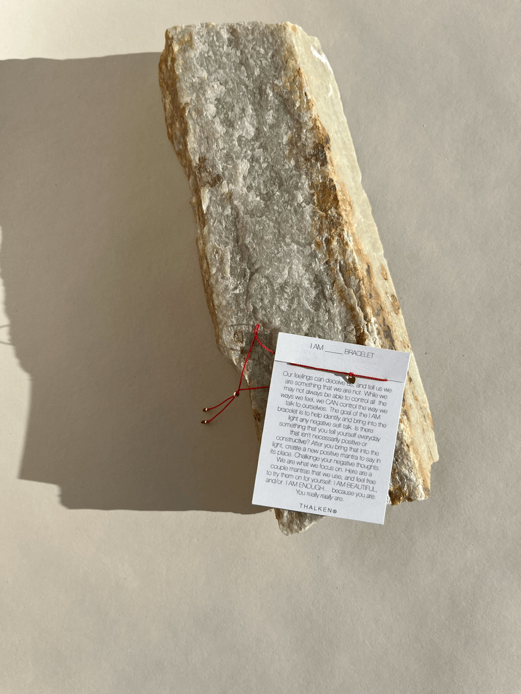 Pulled-back shot of the Thalken I AM bracelet on mantra card resting on a rock surface featuring a thin red cord