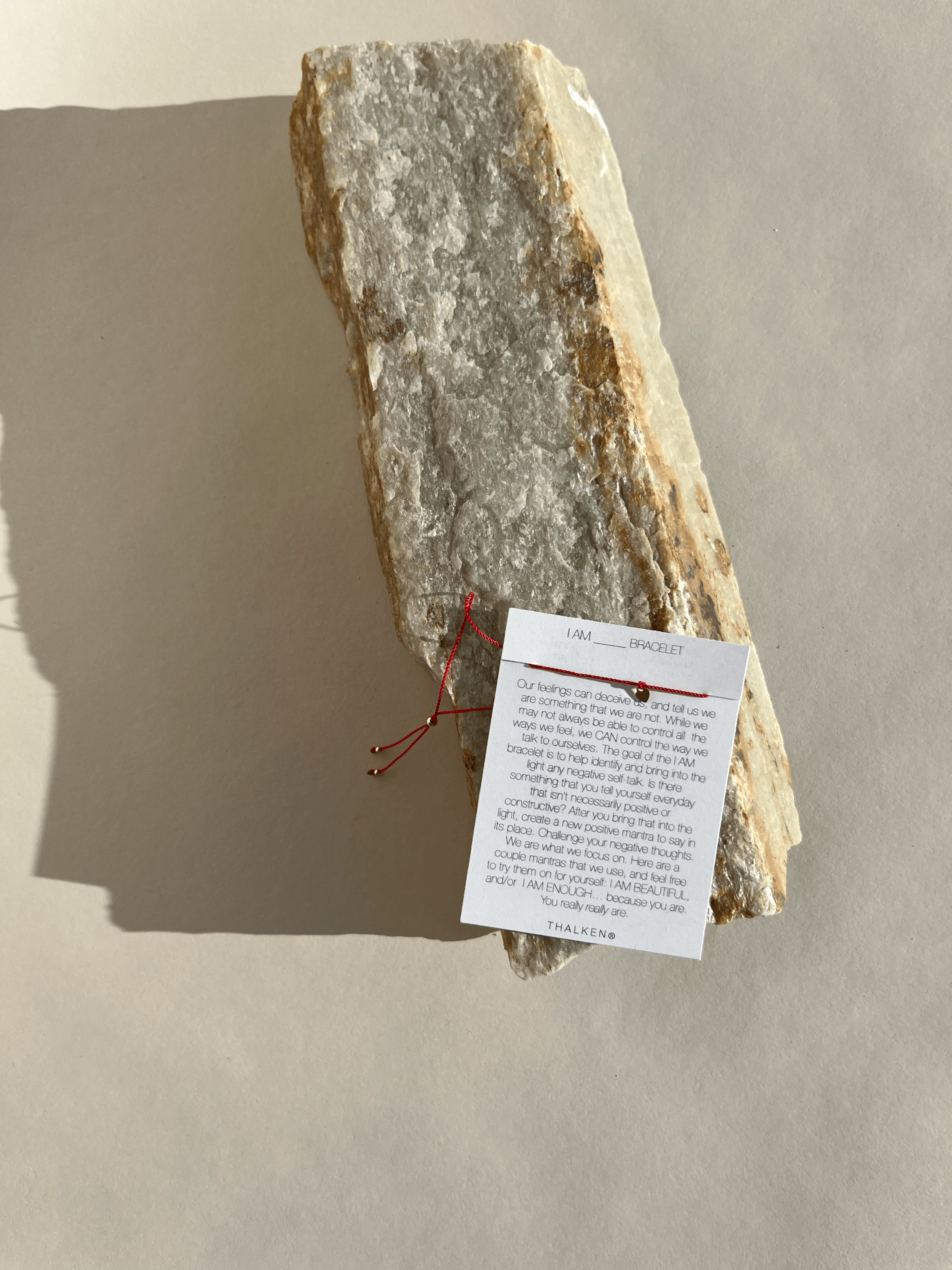 Pulled-back shot of the Thalken I AM bracelet on mantra card resting on a rock surface featuring a thin red cord