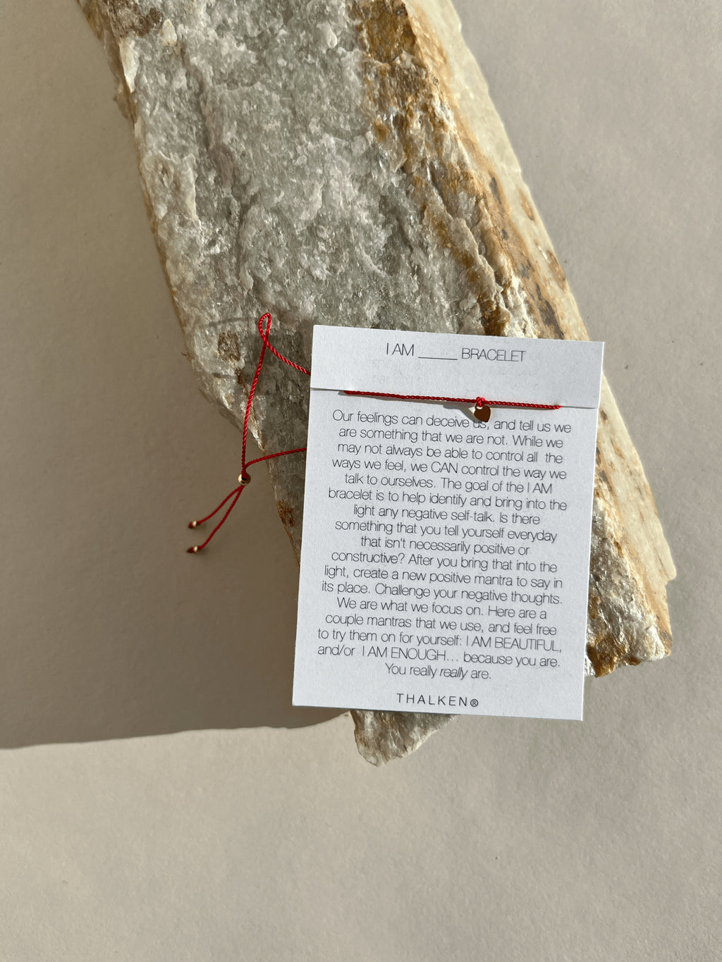 Thalken® I AM bracelet displayed on its mantra card, placed on a natural rock surface, featuring a thin red cord
