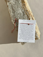 Thalken® I AM bracelet displayed on its mantra card, placed on a natural rock surface, featuring a thin red cord