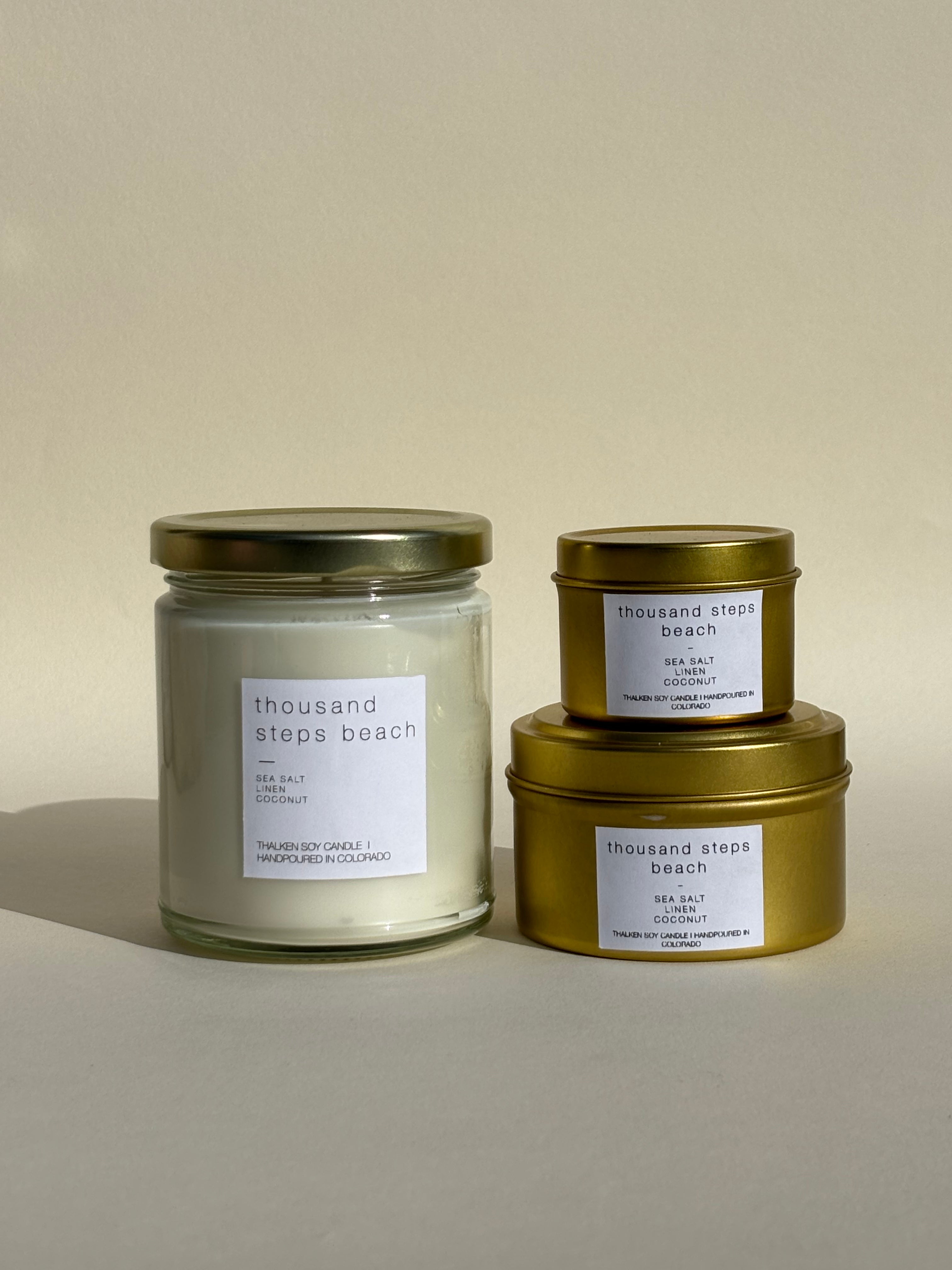 Photo of Thalken’s Thousand Steps Beach candle shown in three vessel options — sleek gold tins (2oz and 6oz) and a soft-glow glass jar (8oz). Bright and sunny scent profile with notes of sea salt, linen, and coconut.