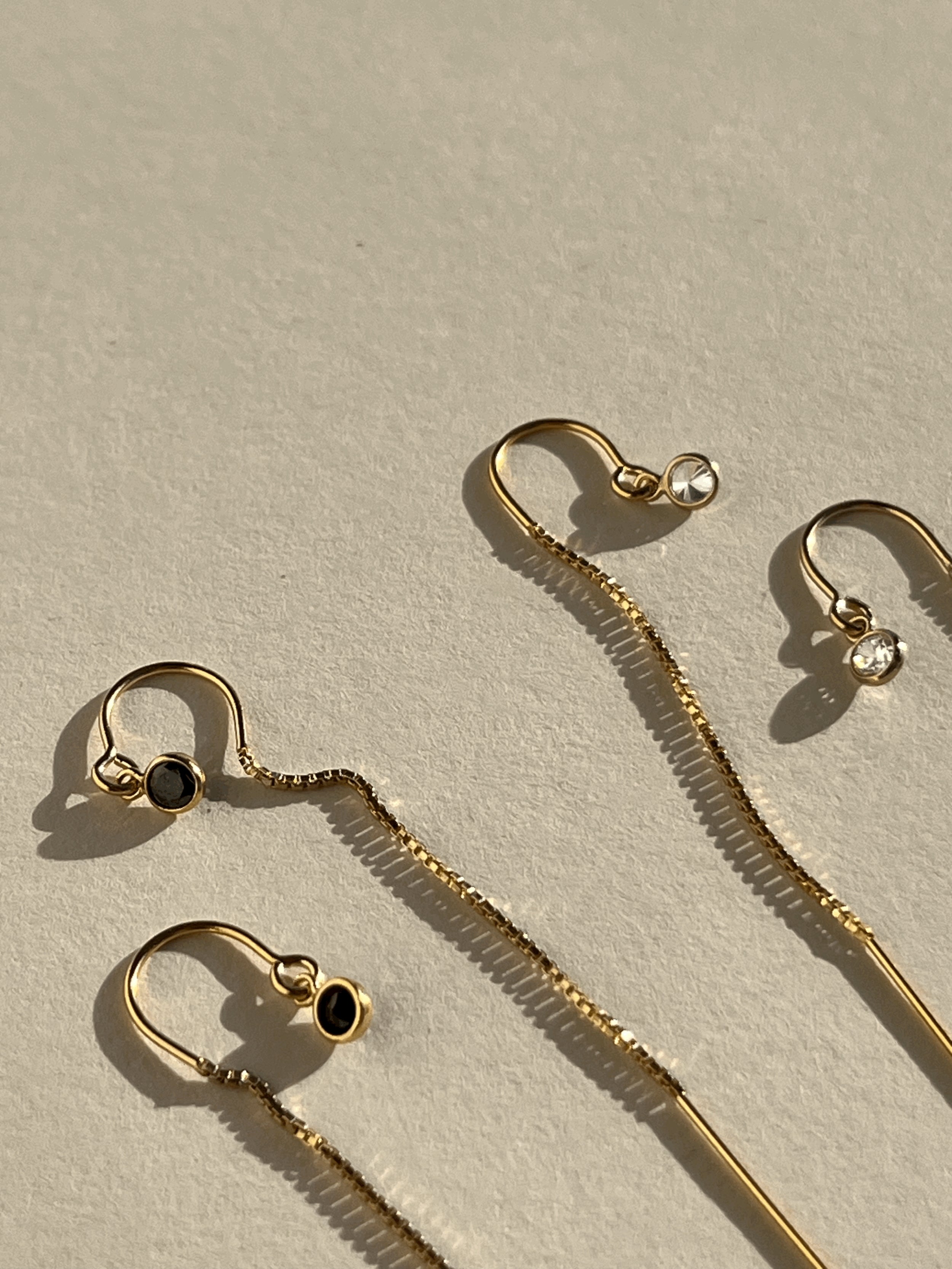 Close-up of wo pairs of Thalken’s yellow gold-filled CZ bezel threader earrings displayed on a neutral background, one with black cubic zirconia and one with clear cubic zirconia