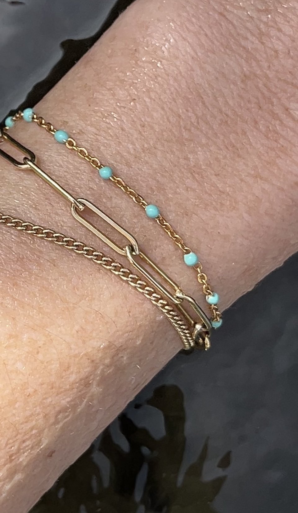 Close-up of model’s wrist in water wearing a yellow gold-filled bracelet stack featuring Thalken® Large Link Bracelet, turquoise enamel bead bracelet, and Perfect Curb Chain Bracelet