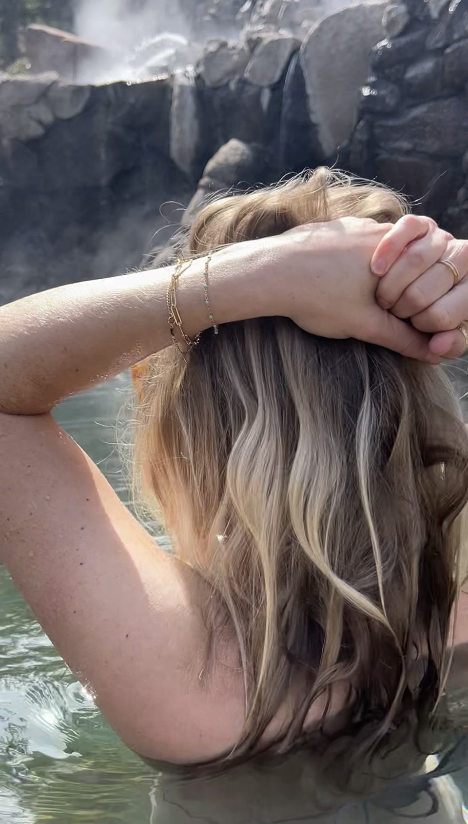 Model in hot springs, photographed from behind, with hands resting on top of flowing hair, showcasing a stack of Thalken® gold-filled bracelets including turquoise enamel bead bracelet