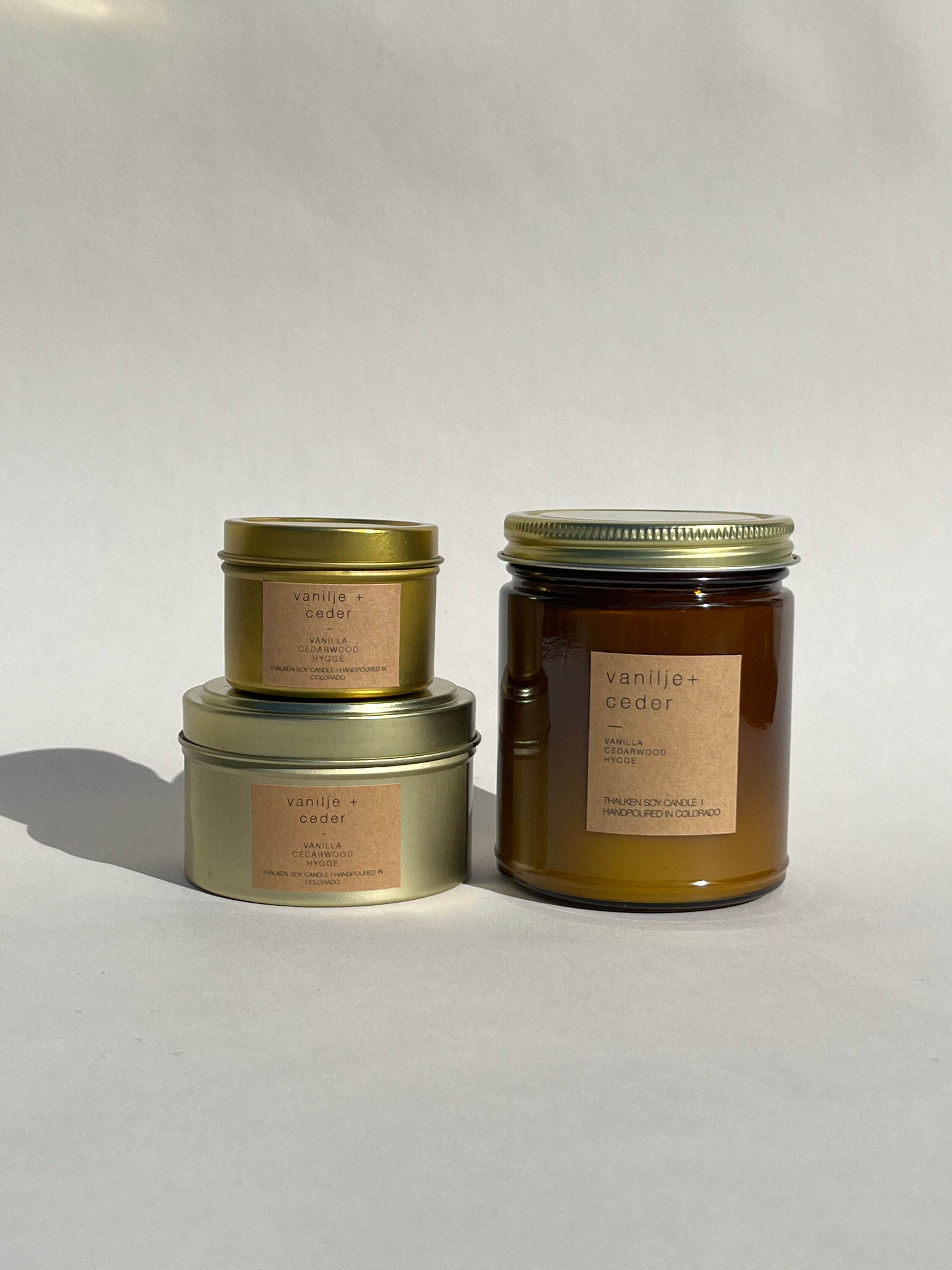 Vanilla and cedar scented candles in three containers: a large brown glass jar, a medium gold tin, and a small gold tin