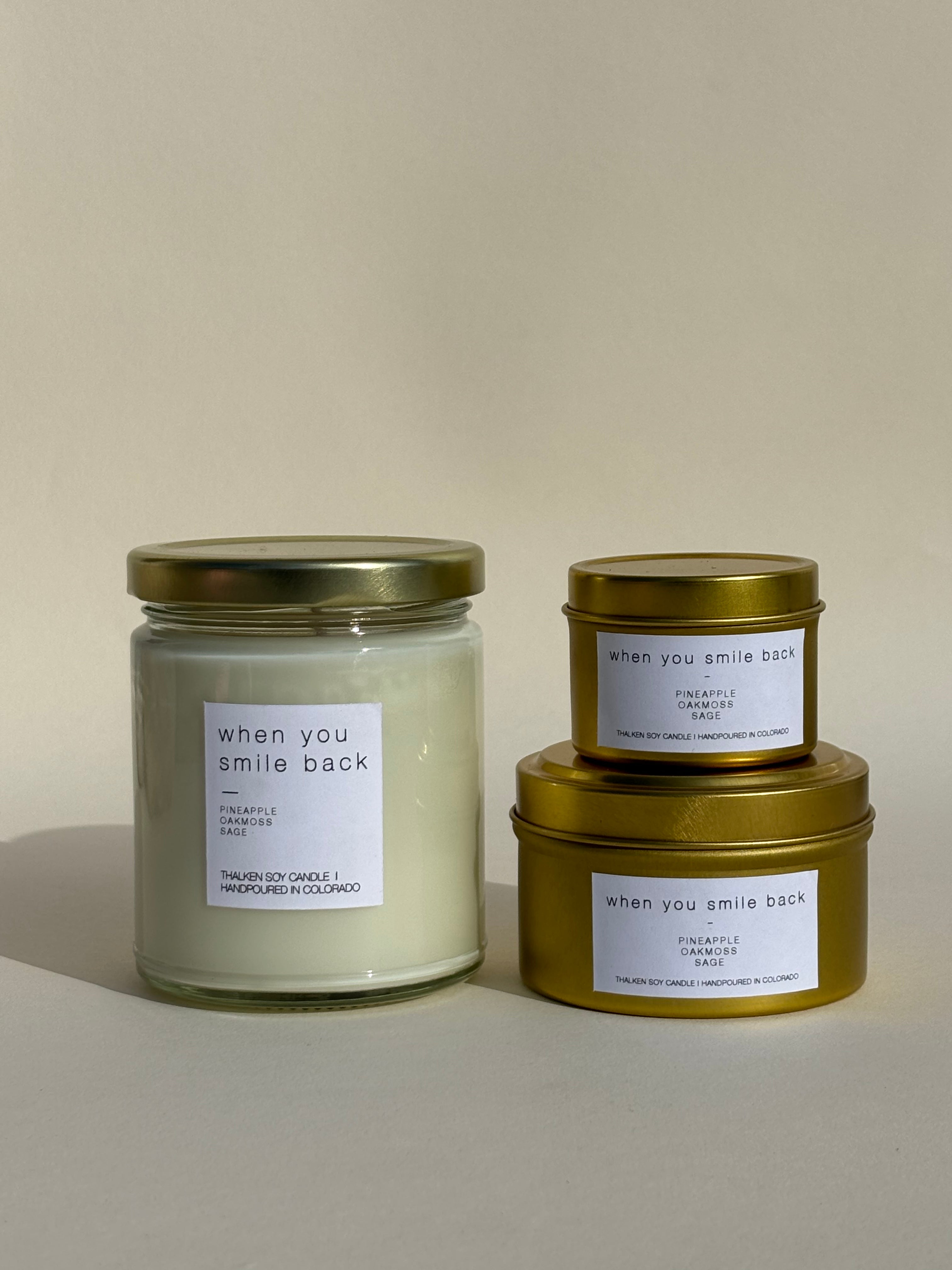 Photo of Thalken’s When You Smile Back candle shown in three vessel options — sleek gold tins (2oz and 6oz) and a soft-glow glass jar (8oz). Bright and sunny scent profile with notes of pineapple, oakmoss, and sage.