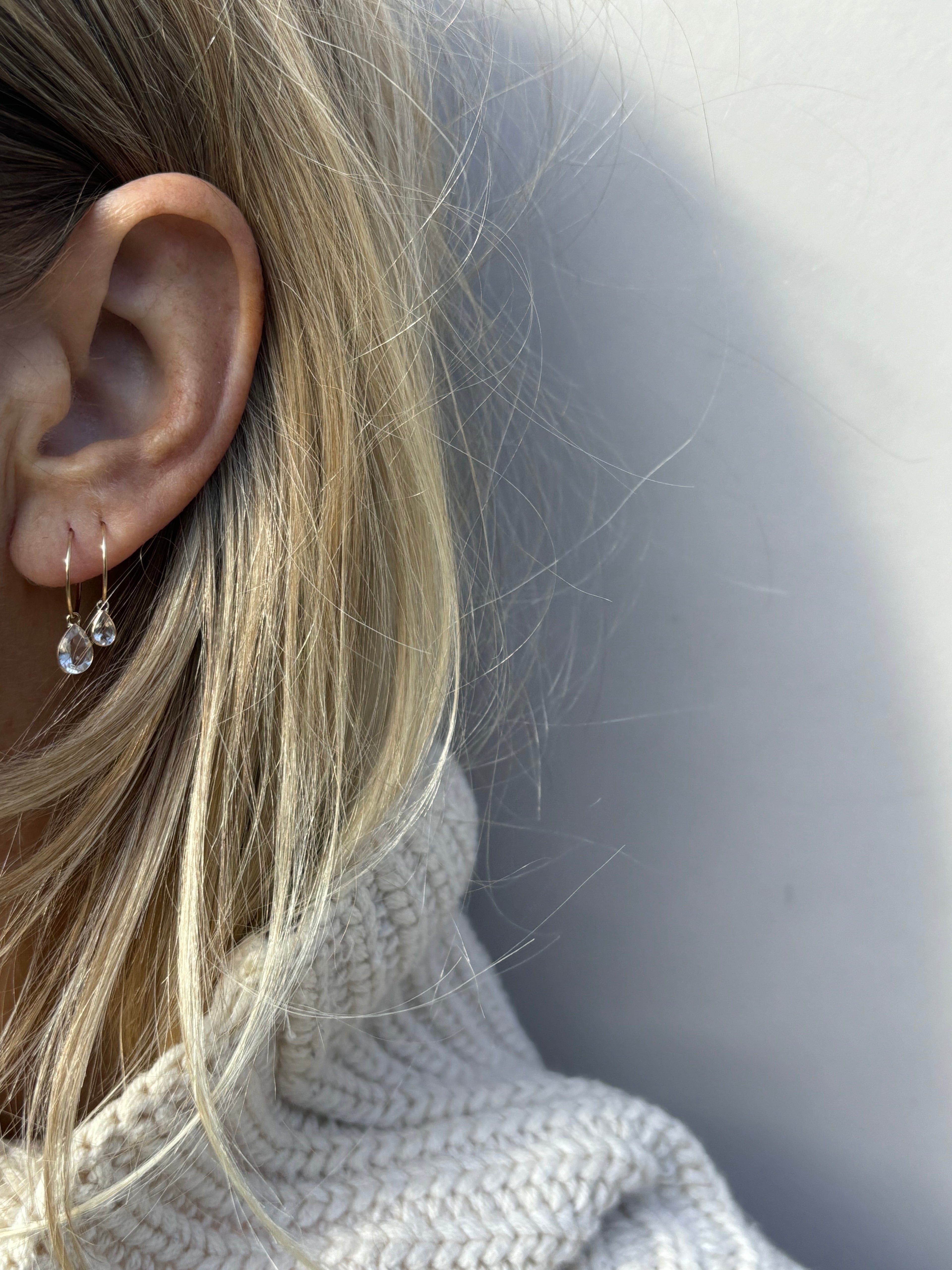 Model wearing Thalken 14k gold 12mm hoop earring with a large pear-shaped white topaz drop in the first ear piercing and a 10mm hoop with a small pear-shaped white topaz drop in the second ear piercing