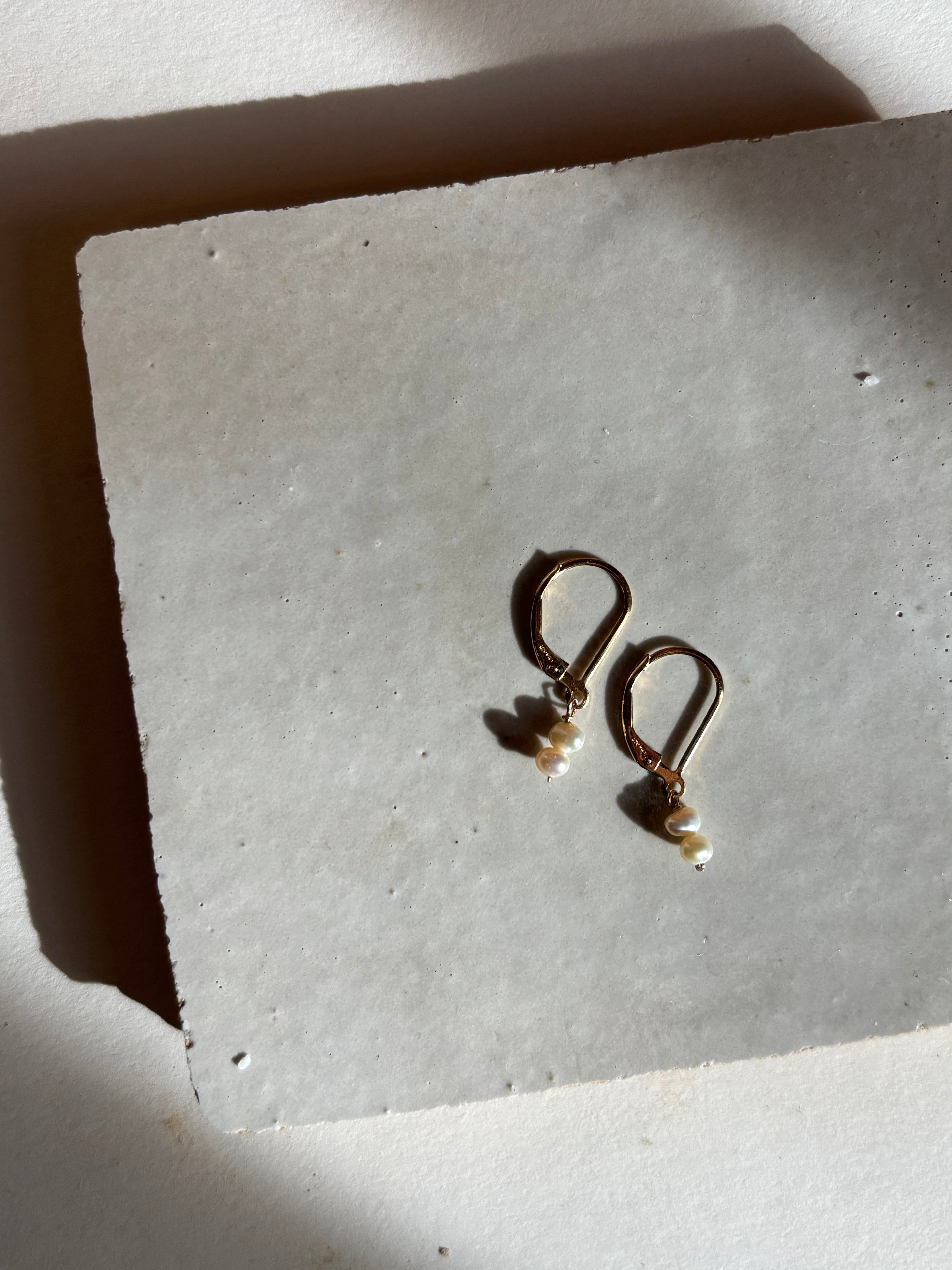 Delicate white double pearl drop Duet Extra Small earrings on an organic surface with warm, moody lighting showing the full earring design and pearl detail.