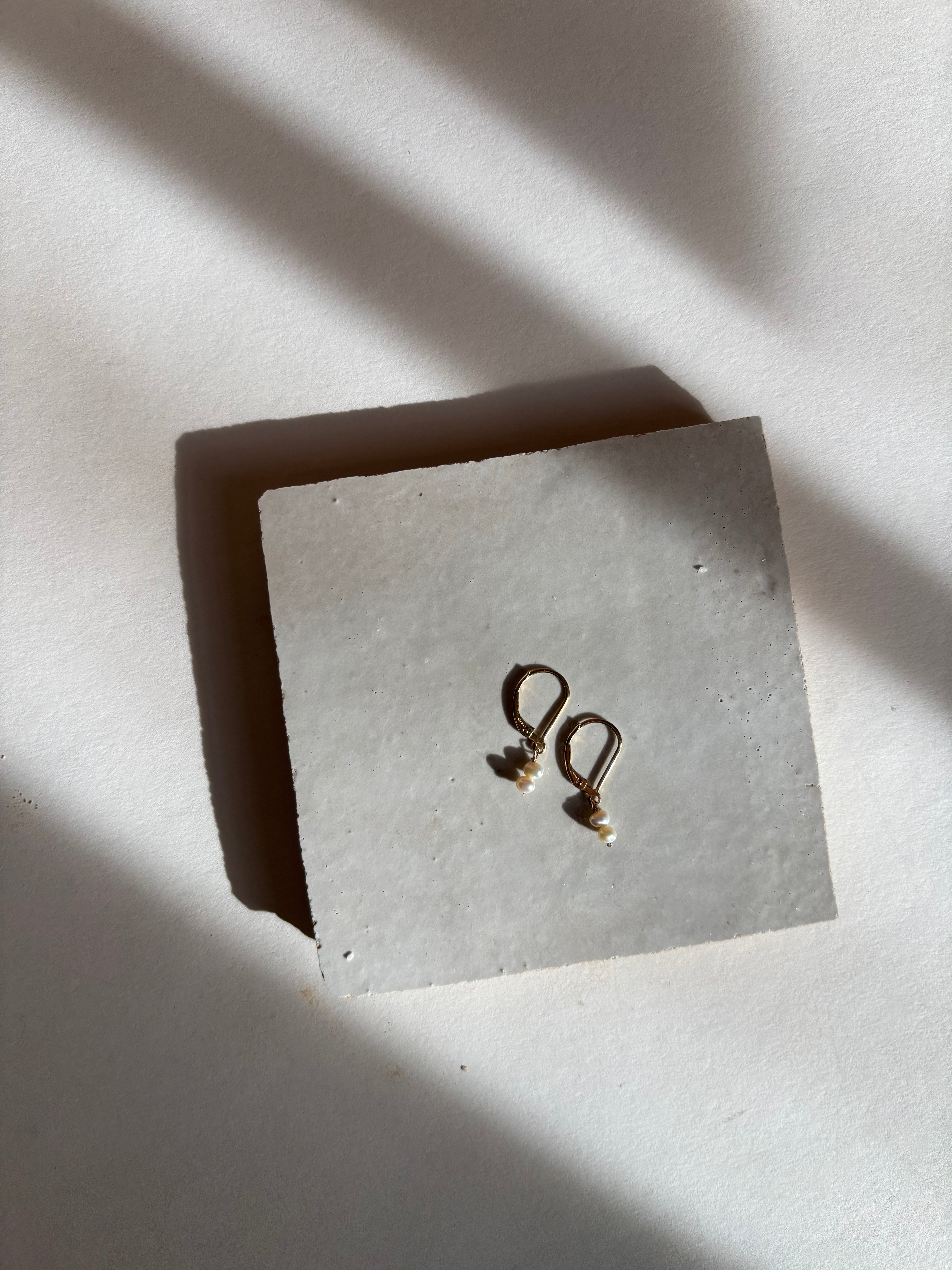 Delicate white double-pearl drop Duet Extra Small earrings on an organic surface with warm, moody lighting, showing the full earring design and pearl details. Bird’s-eye view shot.