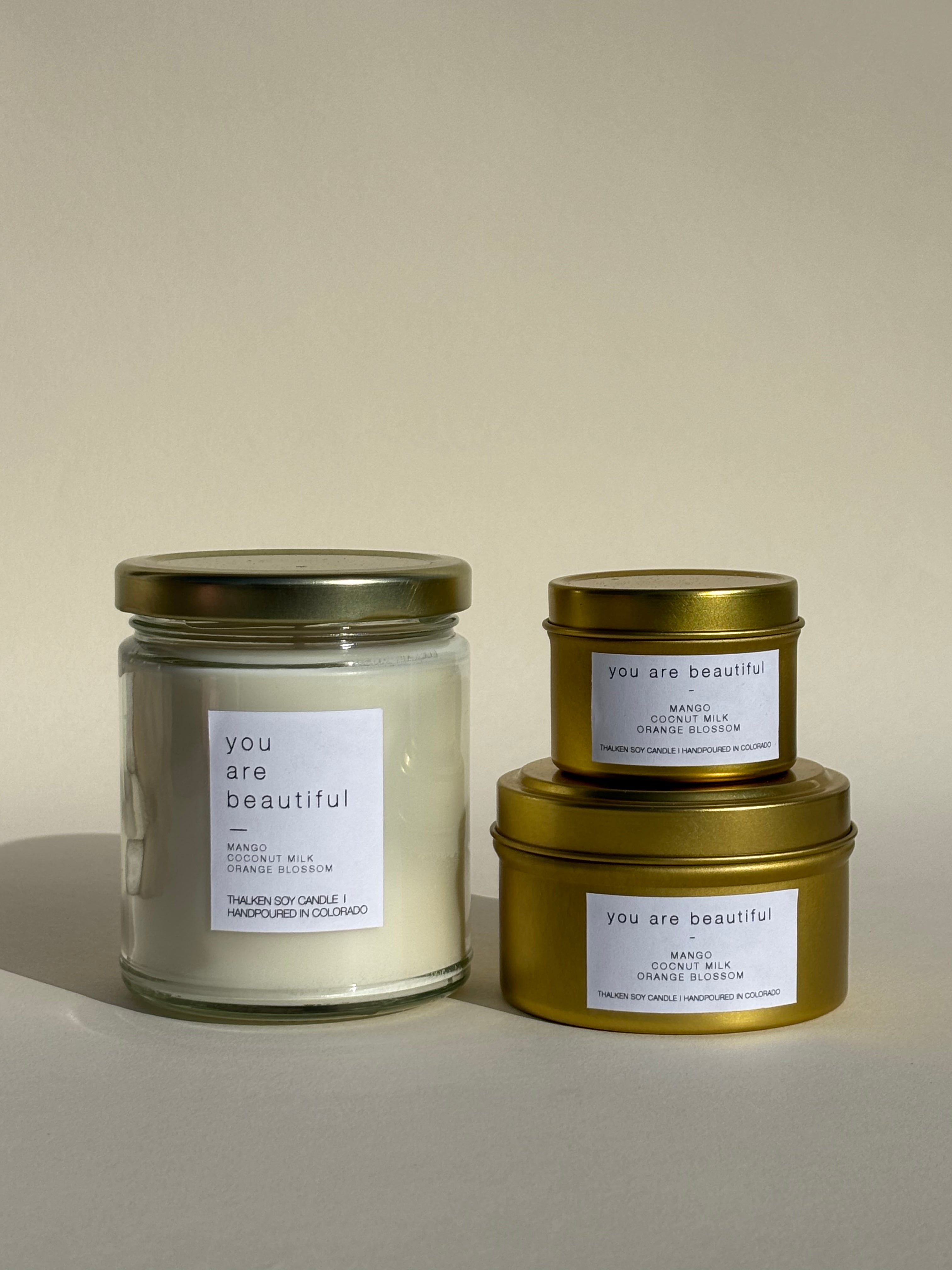 Photo of Thalken’s You Are Beautiful candle shown in three vessel options — sleek gold tins (2oz and 6oz) and a soft-glow glass jar (8oz). Bright and sunny scent profile with notes of white tea, mango, and coconut.