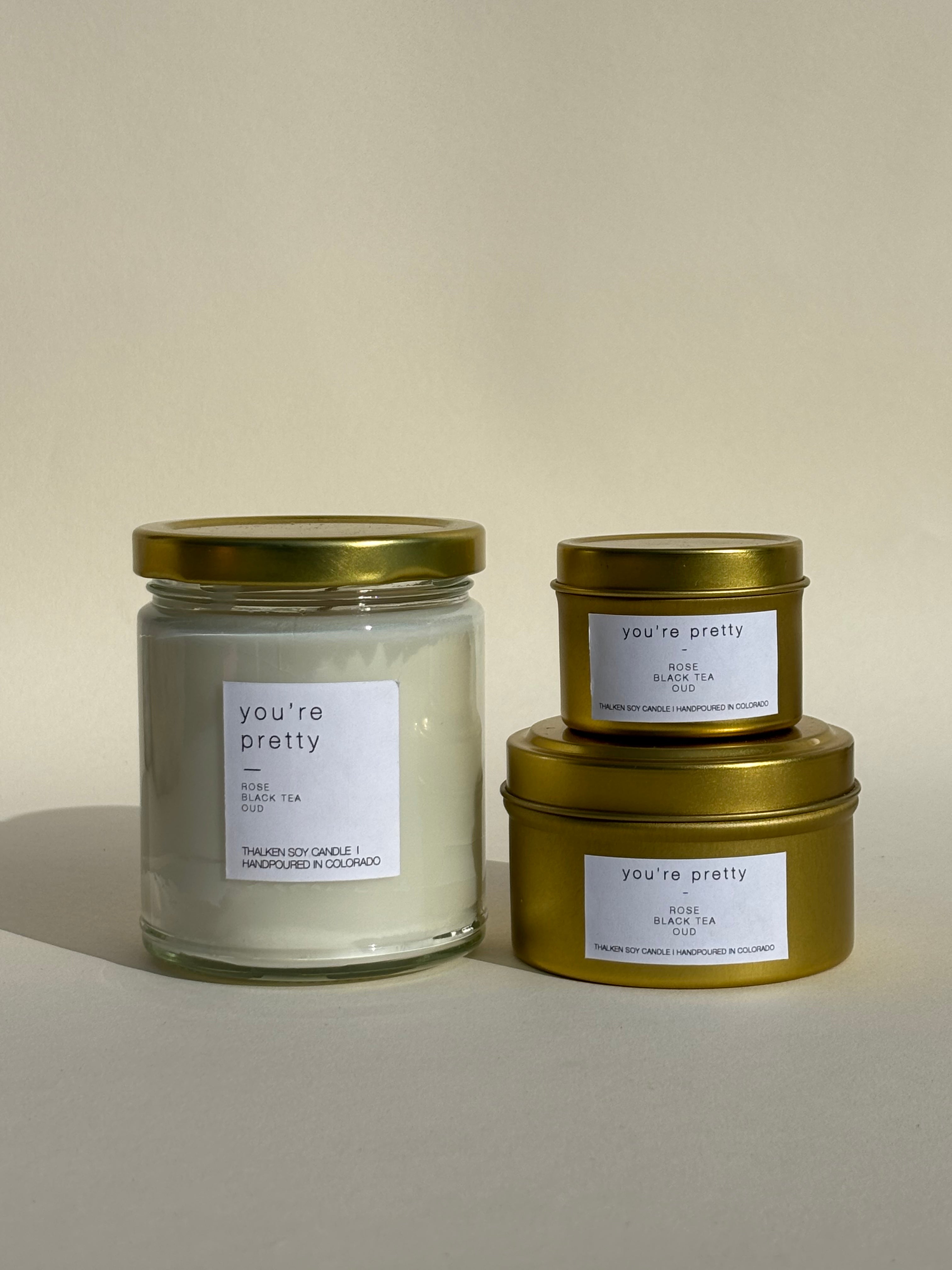 Photo of Thalken’s You're Pretty candle shown in three vessel options — sleek gold tins (2oz and 6oz) and a soft-glow glass jar (8oz). Bright and sunny scent profile with notes of rose, black tea, oud.